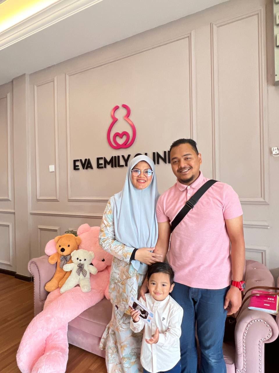 A family of three standing in front of a pink sign that says 'Eva Emily Clinic' with a whale-shaped logo. They are inside a room with a pink plush teddy bear and stuffed animal on a pink couch. The woman is wearing a hijab and glasses, the man is in a pink shirt with a black shoulder strap, and the young boy is holding a card.
