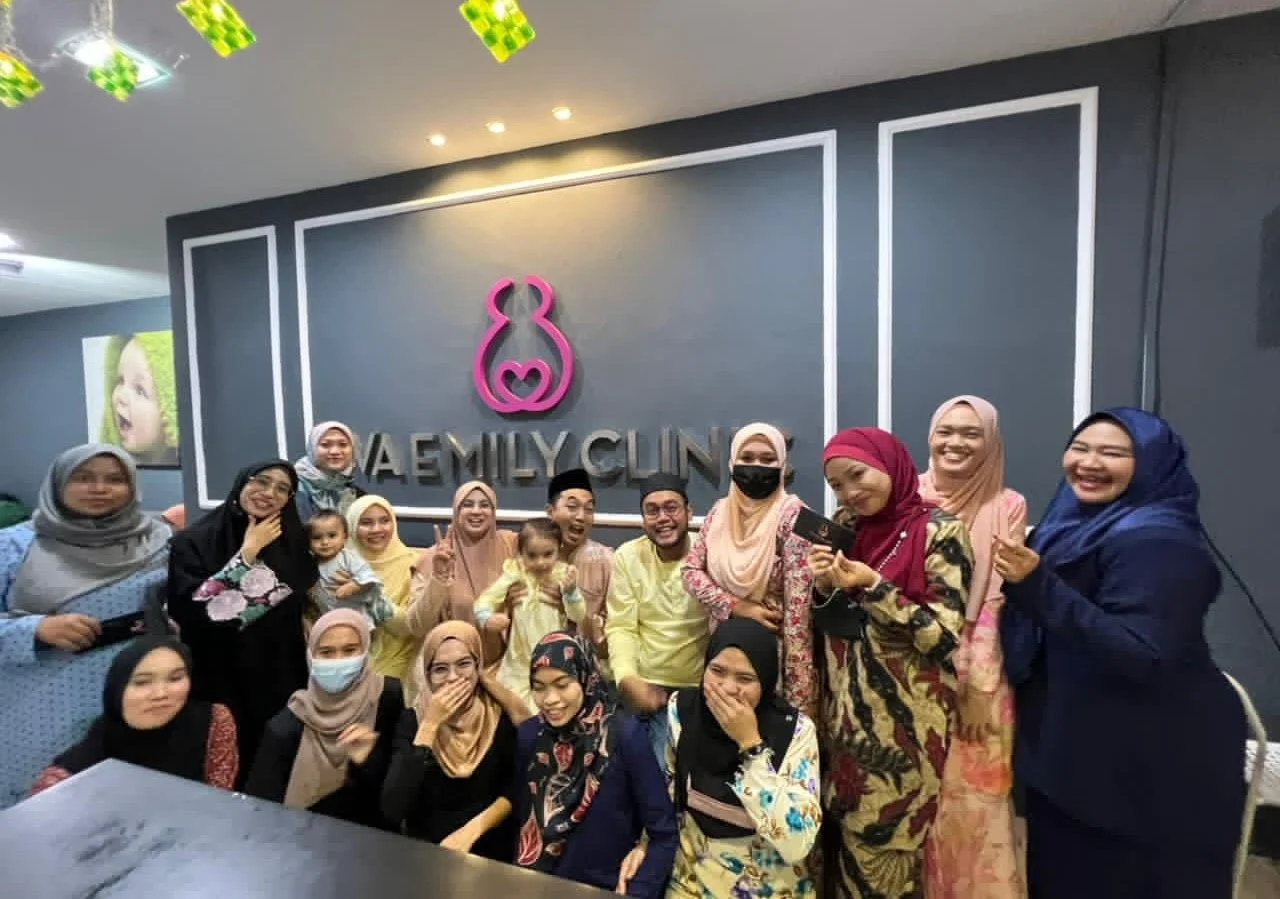 Group of people, mostly women wearing colorful hijabs, gathered inside a clinic with a sign that says 'EVA EMILY CLINIC,' posing and smiling for a photo.