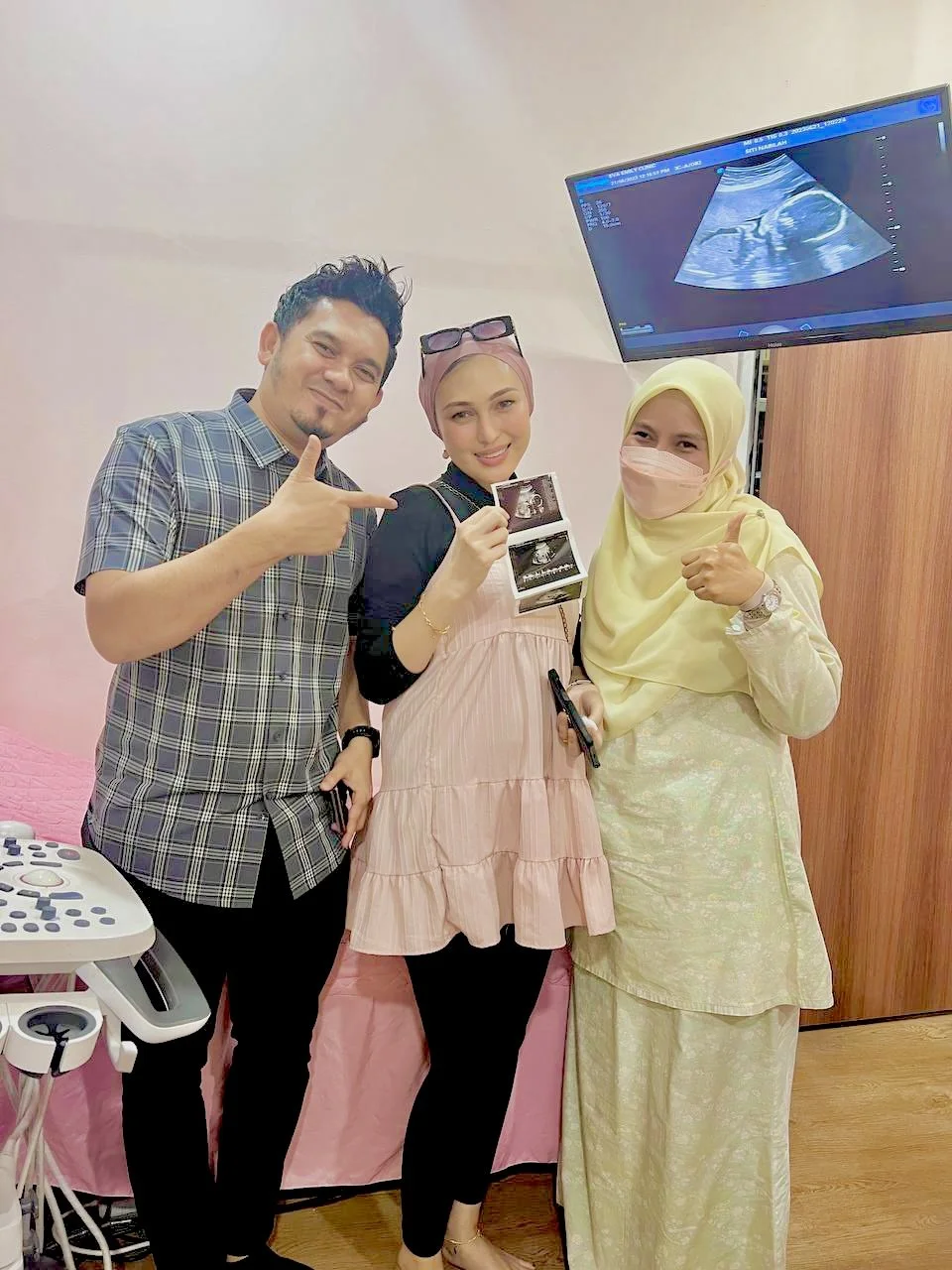 Three people smiling and holding ultrasound images in a medical setting, with an ultrasound machine and a monitor displaying an ultrasound scan.