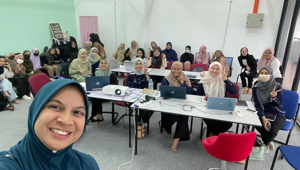 Group of women in a conference room taking a selfie, some wearing face masks, with laptops and a projector on the table.