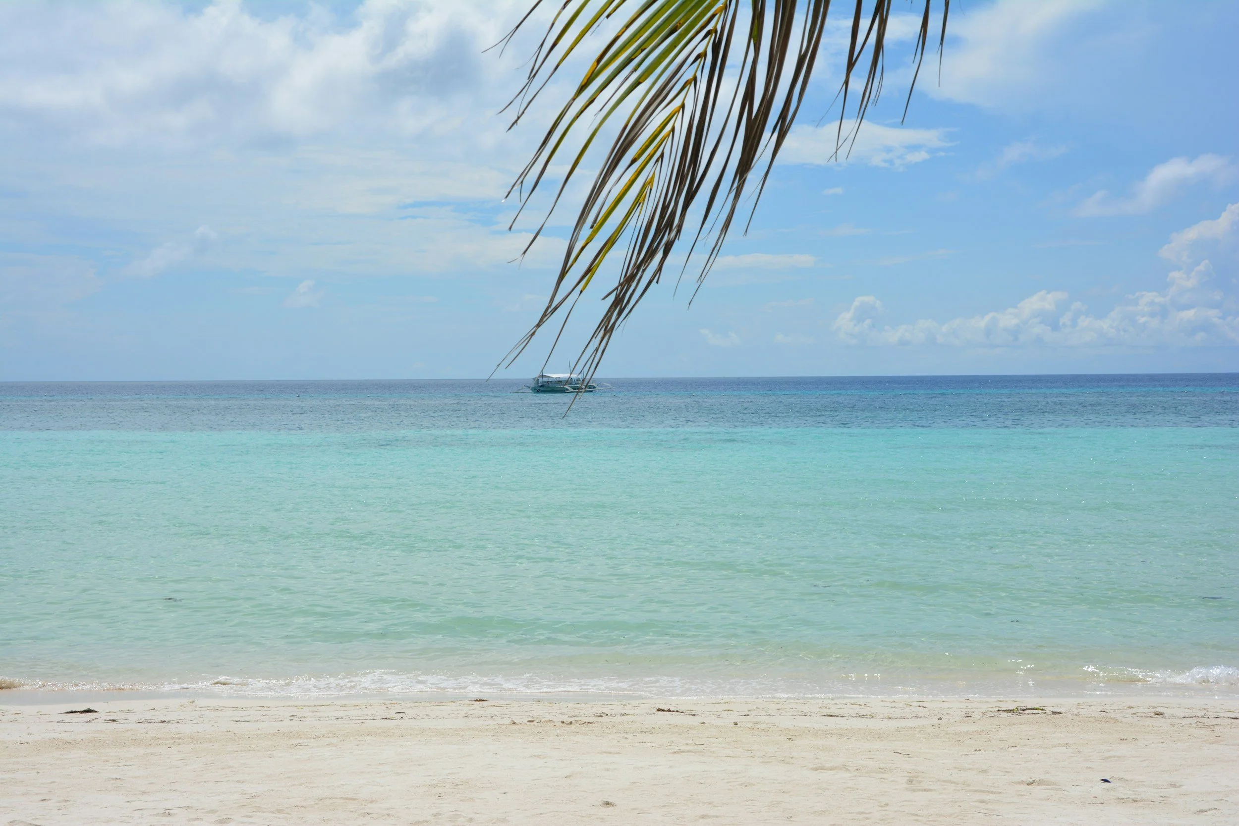 Tropical beach with white sand, turquoise water, blue sky with a few clouds, a yacht in the distance, and palm tree leaves hanging down from the top.
