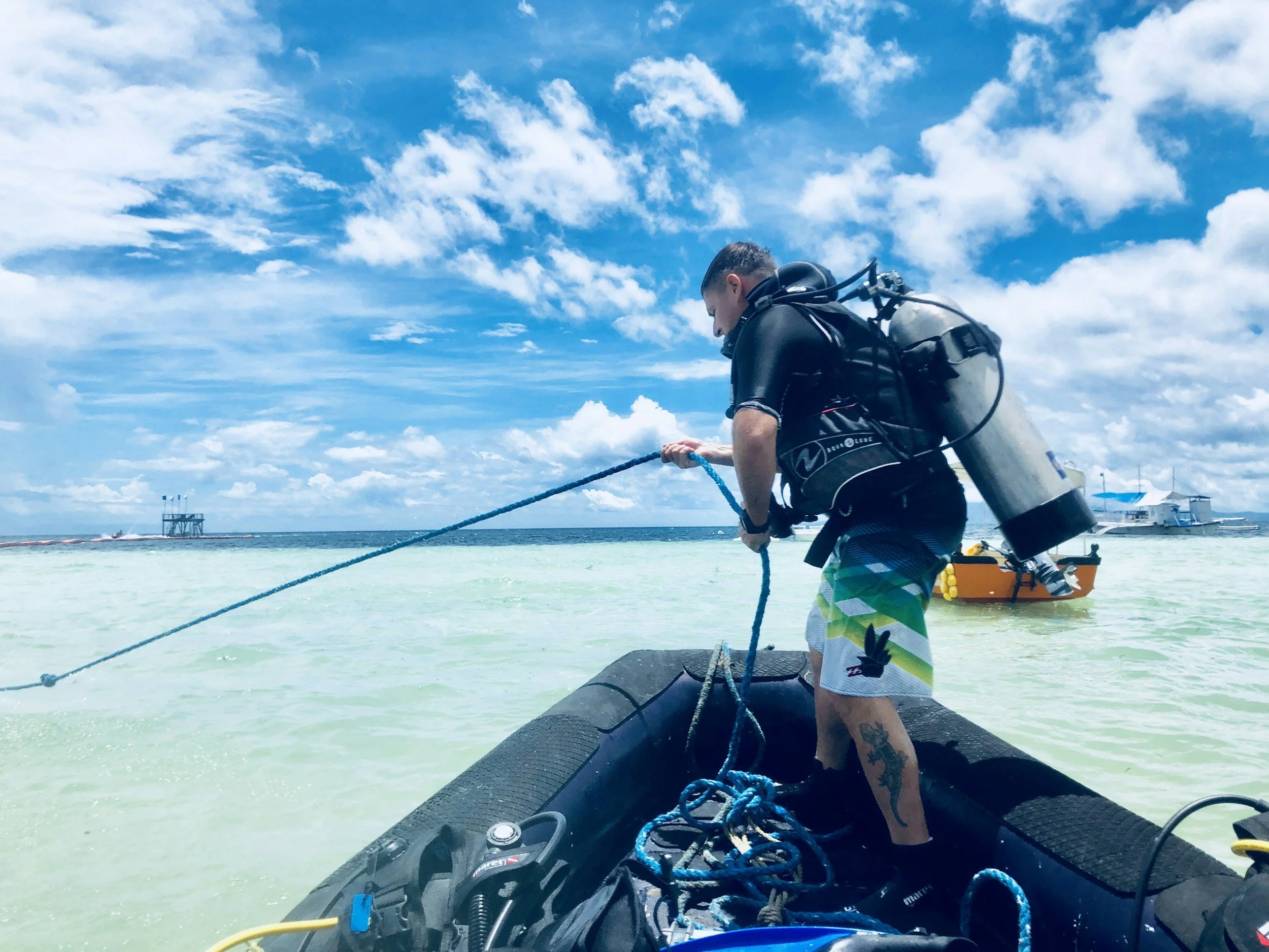 Man in wetsuit with scuba tank on back on inflatable boat, holding a rope in clear tropical water with a partly cloudy sky.