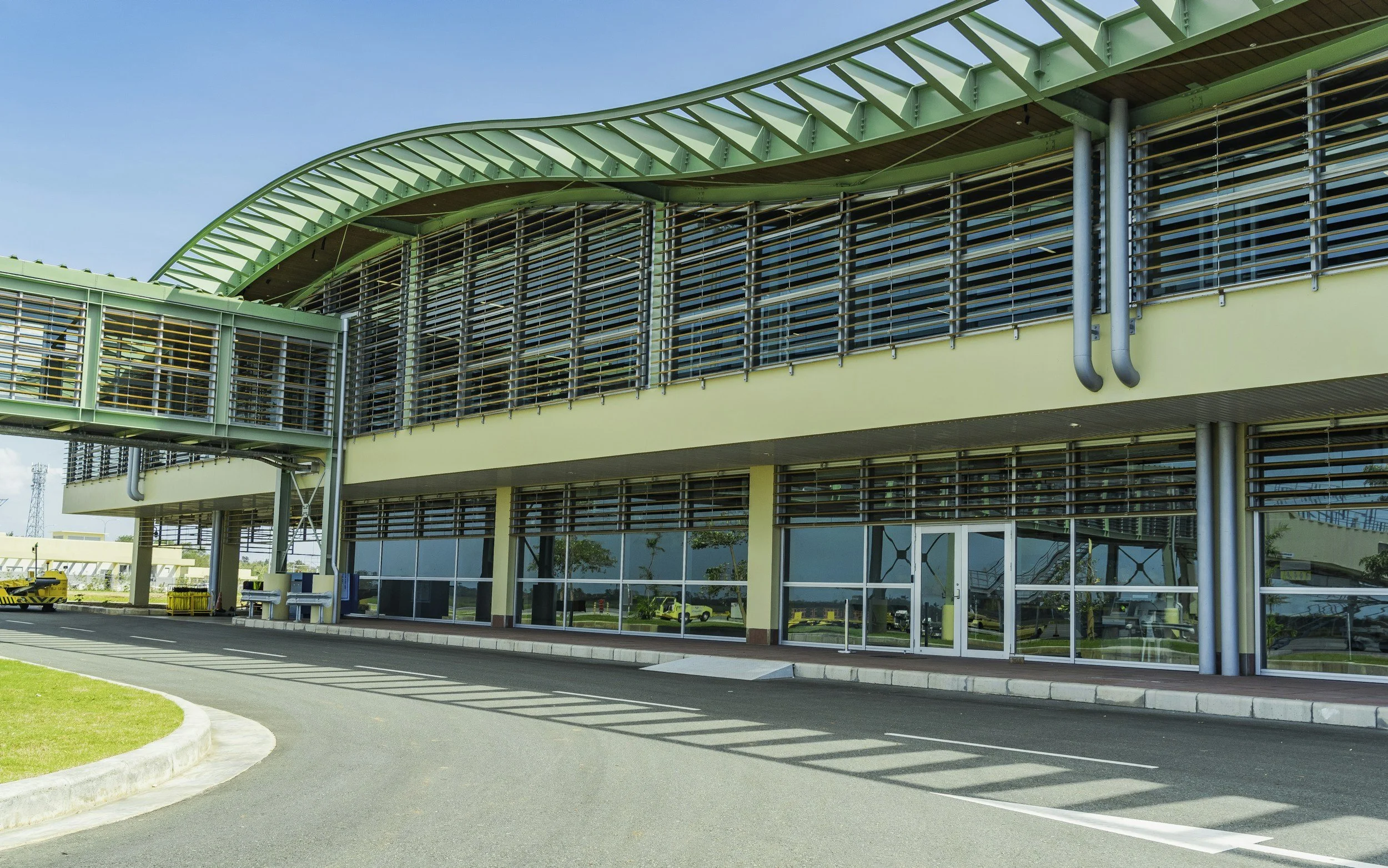 Modern airport terminal building with glass windows, metal structures, and an overhanging green roof, with a curved driveway in front.