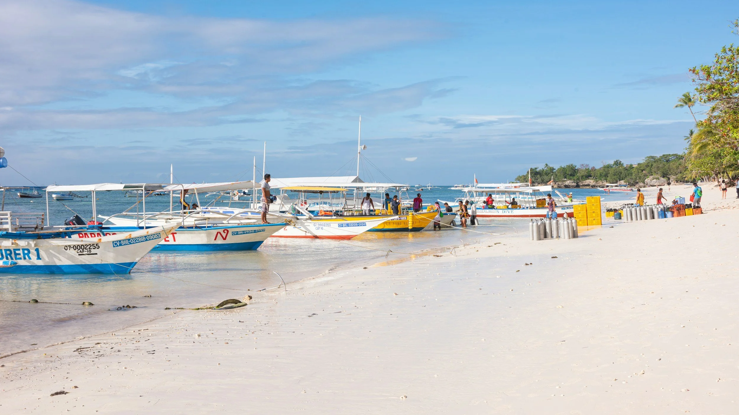 Boats docked at a sandy beach with people boarding and swimming, clear blue sky, and tropical trees in the background.