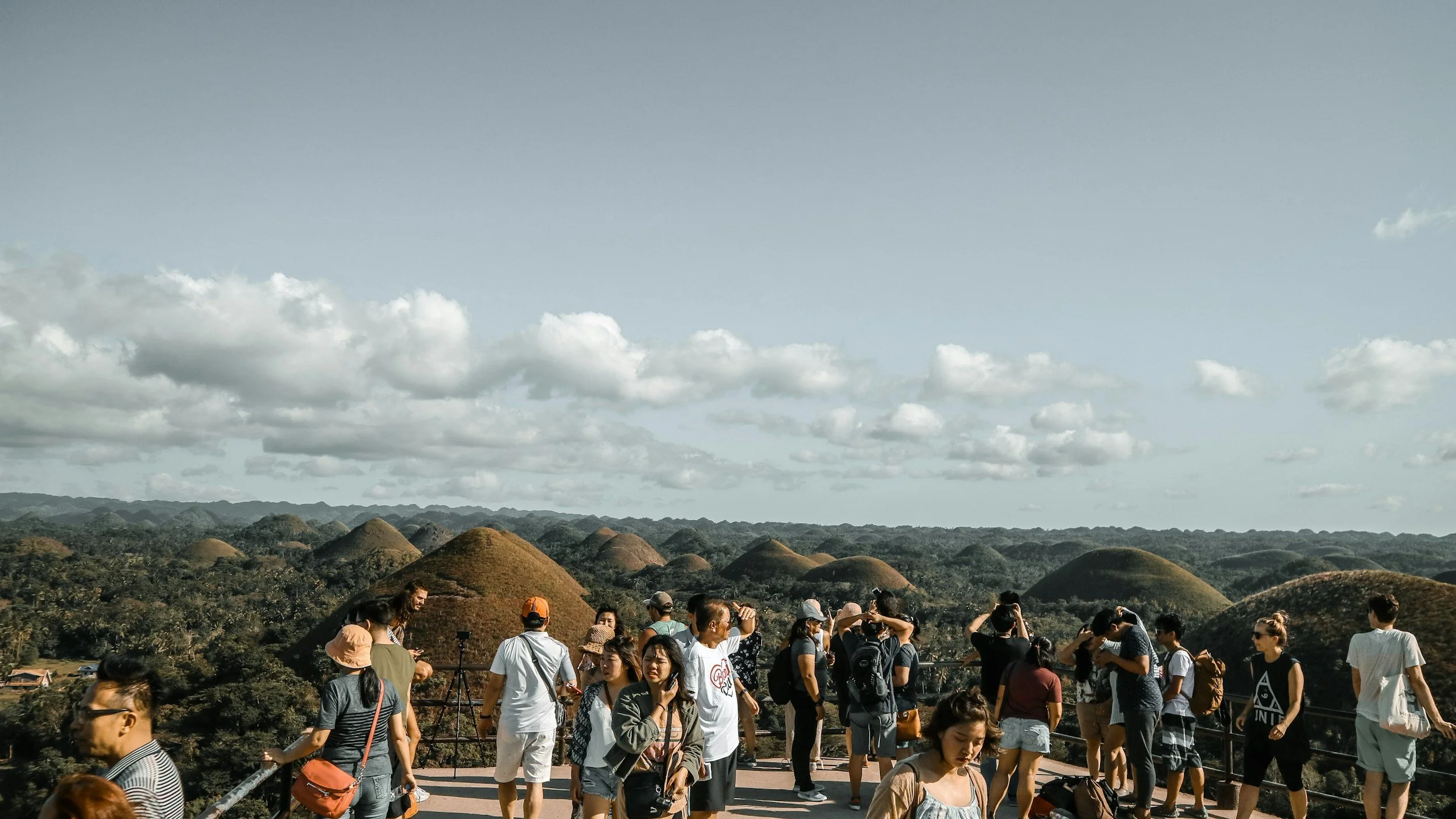 Group of tourists and visitors on a viewpoint overlooking the colorful Chocolate Hills in the Philippines with rolling hills and blue sky with clouds in the background.