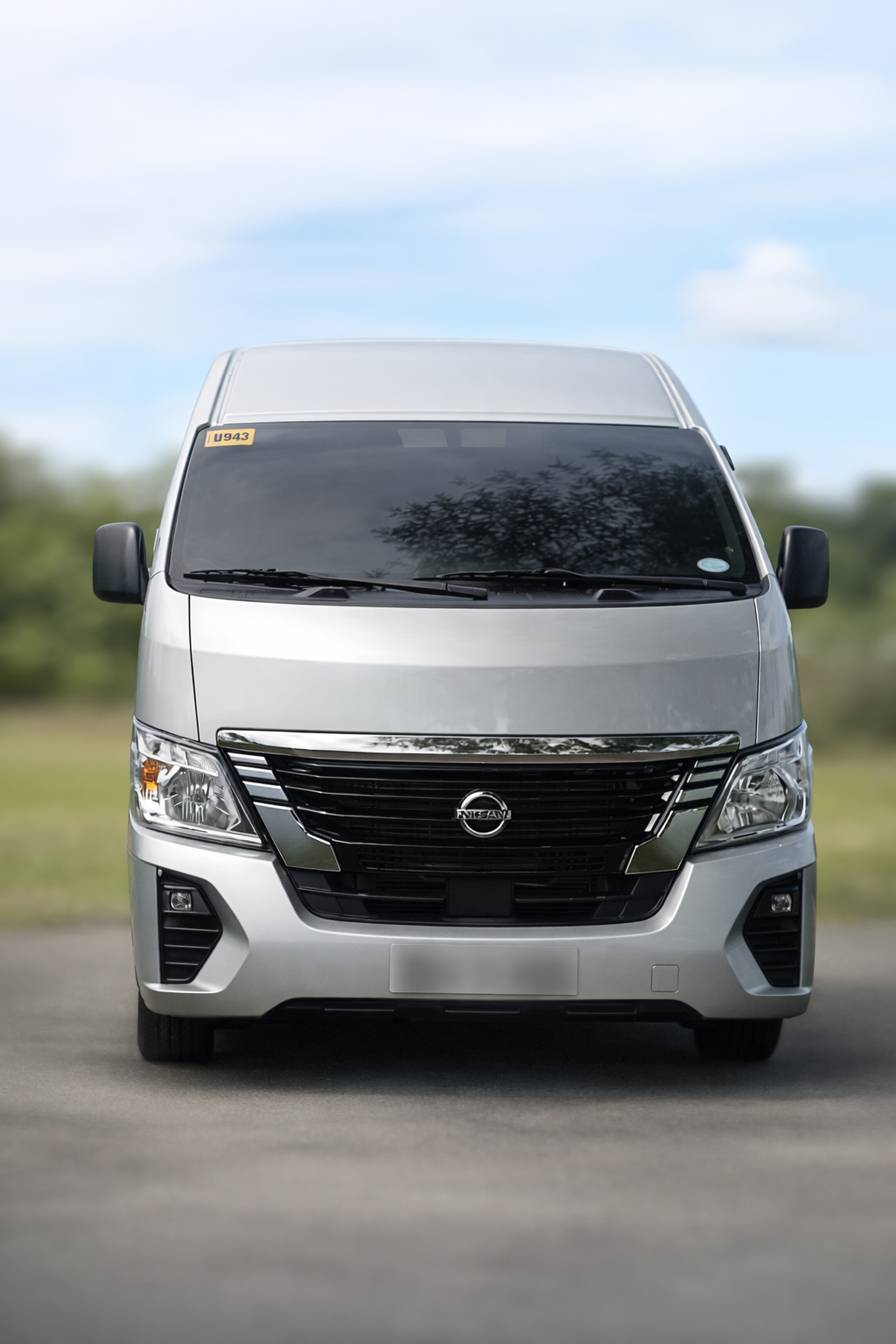Front view of a silver Nissan commercial van parked on a road with trees and a cloudy sky in the background.