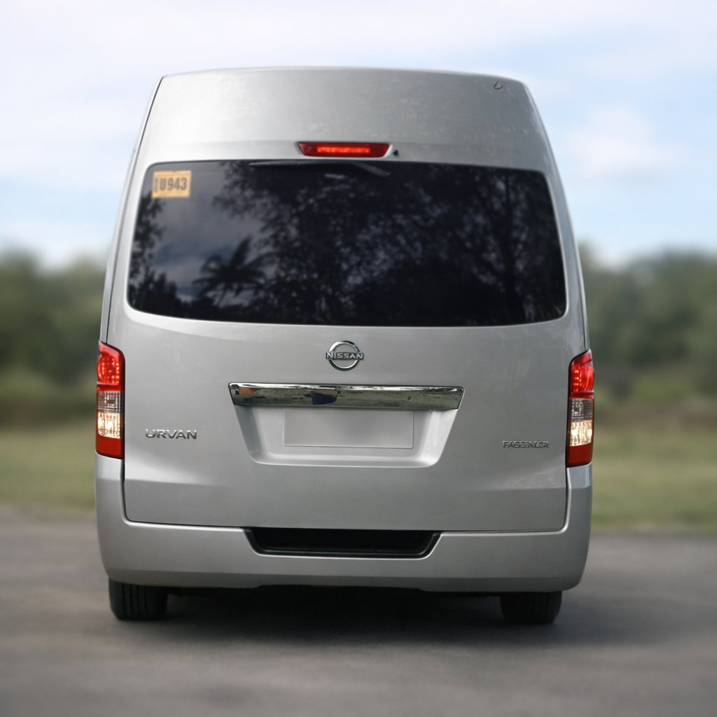 Silver Nissan Urvan passenger van parked outdoors, showing the rear view with tinted windows and a world license plate.