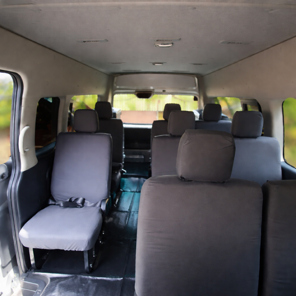 The interior of a van with multiple rows of black seats, facing forward. The van has windows on each side and a flat floor, with a neutral ceiling and walls.