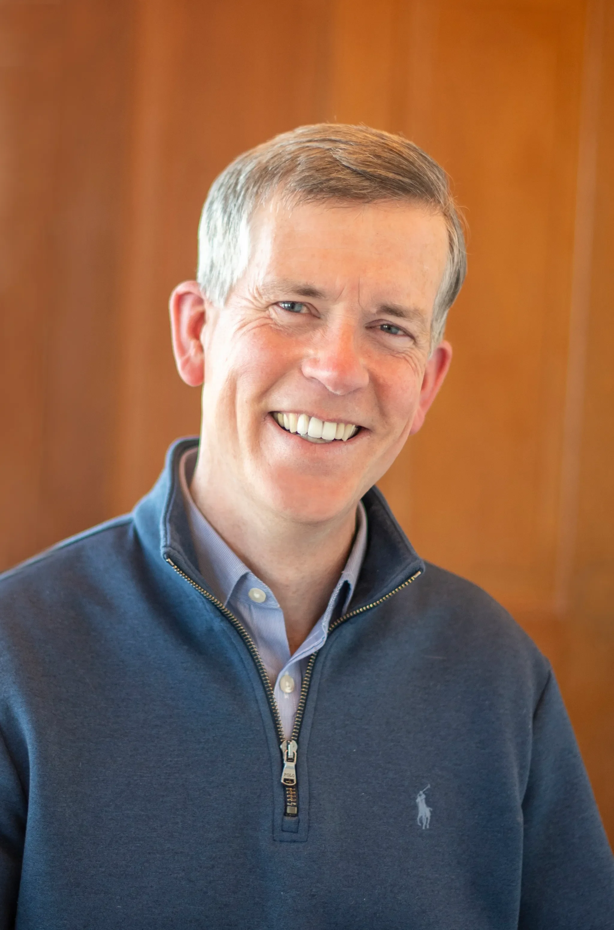 A smiling man with short light brown hair and blue eyes, wearing a blue pullover with a zip, and a collared shirt underneath. The background is blurred with warm brown tones.