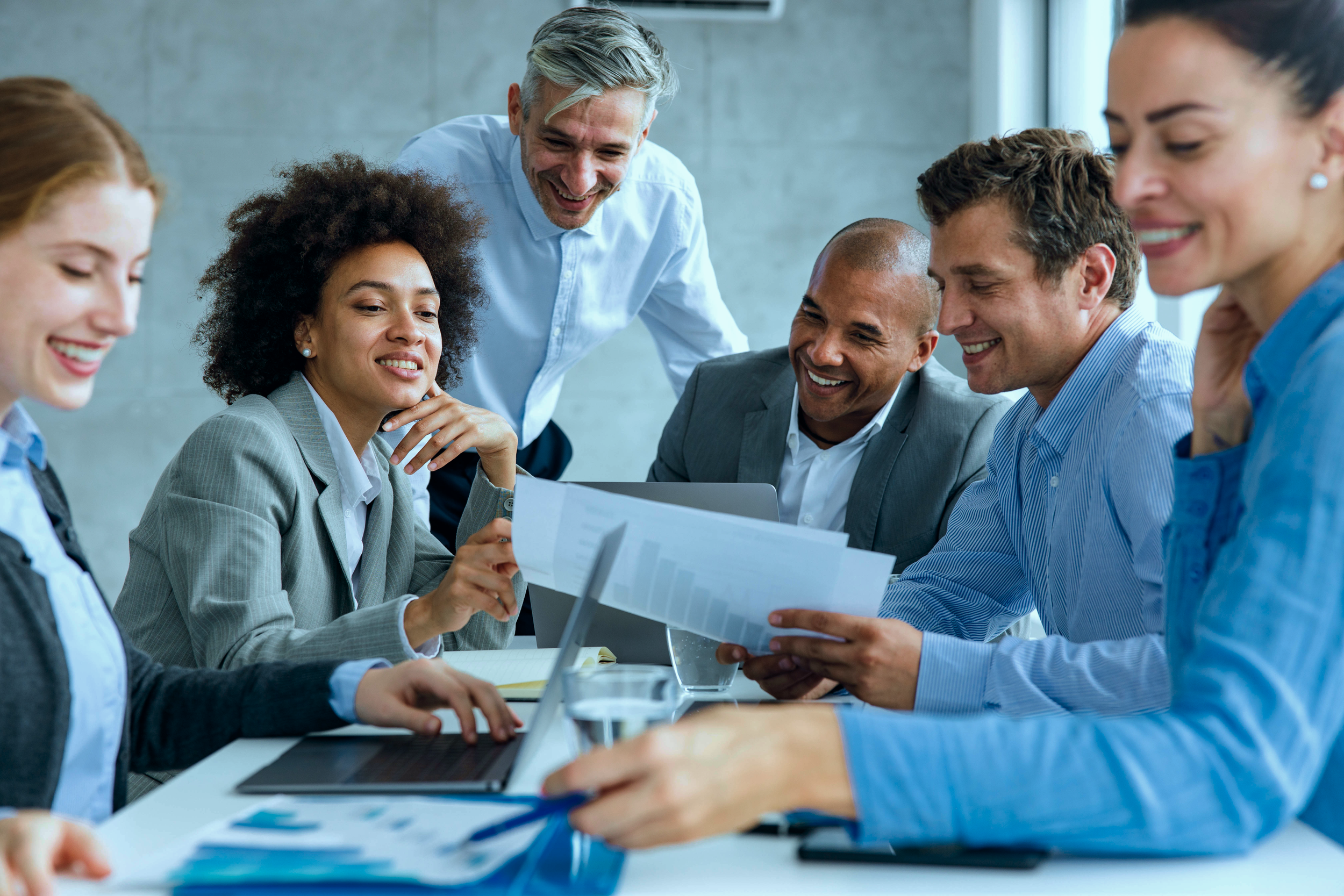 A diverse group of professionals gathered around a table in a meeting room, engaging in a discussion and sharing documents, with a man standing and smiling at the center.