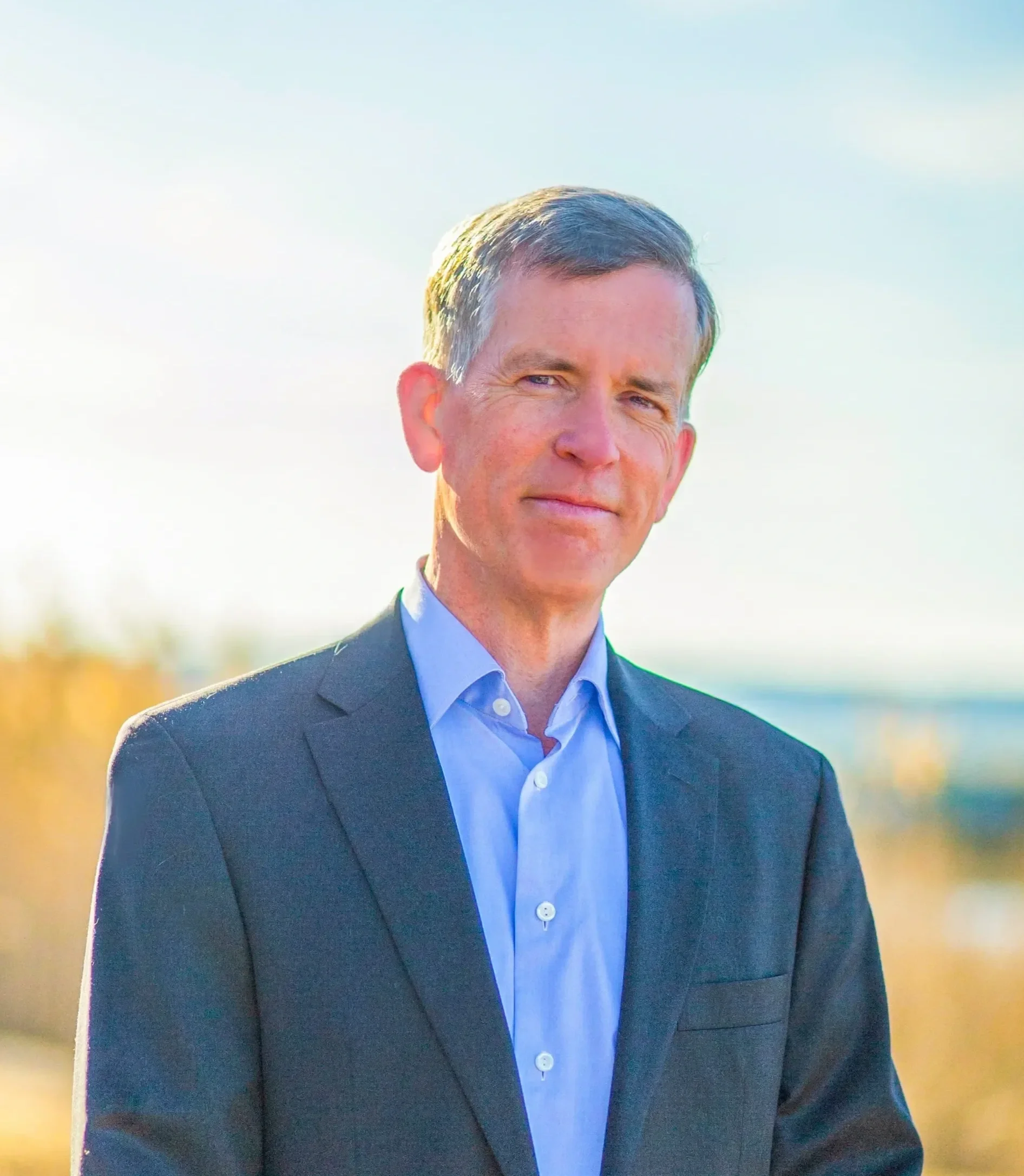 A man with short gray hair and blue eyes, wearing a dark suit jacket and light blue shirt, standing outdoors with a blurred yellow and green background.