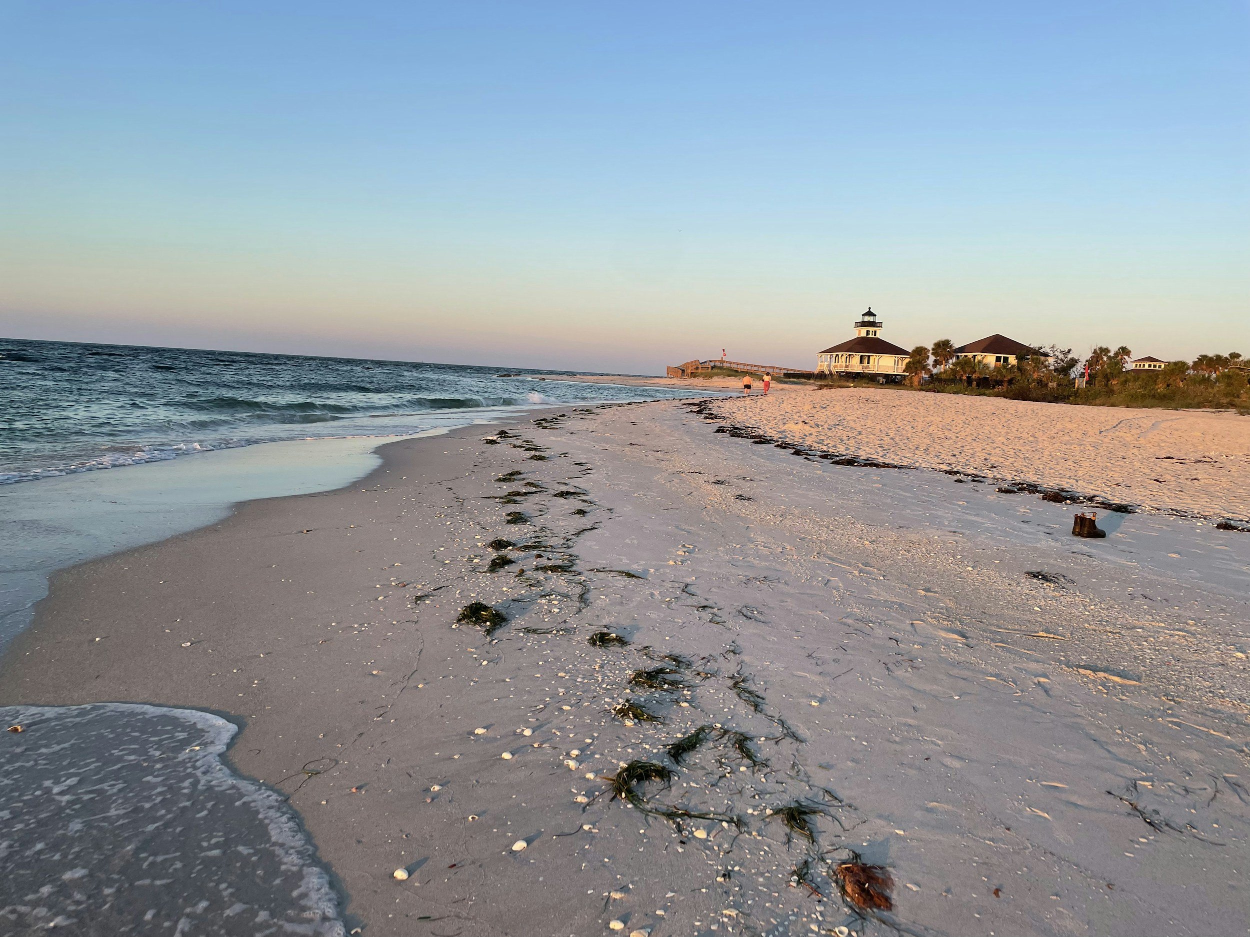 Sunset on a sandy beach with seaweed and shells, buildings and a lighthouse in the background, few people walking along the shore.