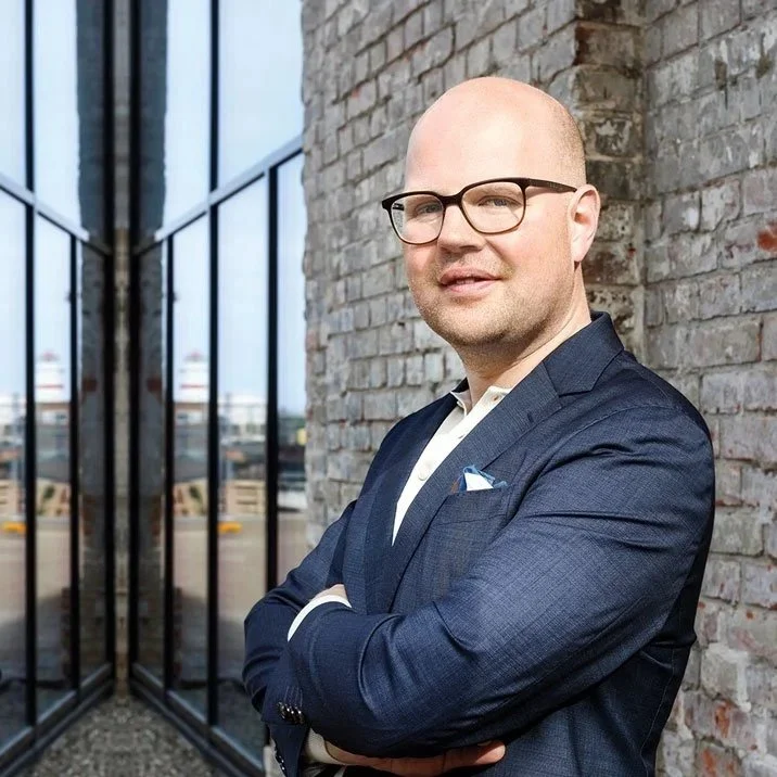 Tobias Sturesson in a navy suit with glasses standing outside near a brick wall, arms crossed, with glass windows reflecting an outdoor scene.