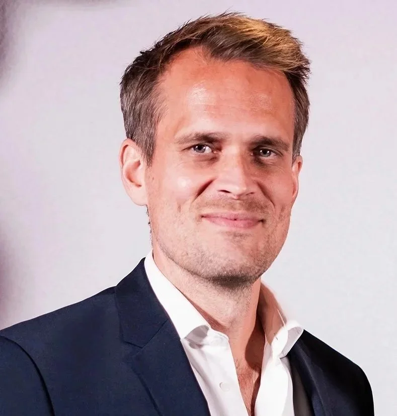 Andreas Almlöf with short light brown hair, wearing a dark suit jacket and white shirt, standing against a plain light background.