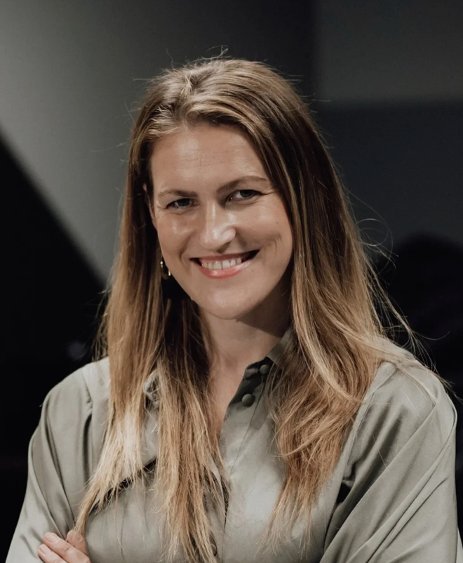 Lena Sturesson with long, wavy, light brown hair smiling at camera, wearing a gray blouse, with a dark background.