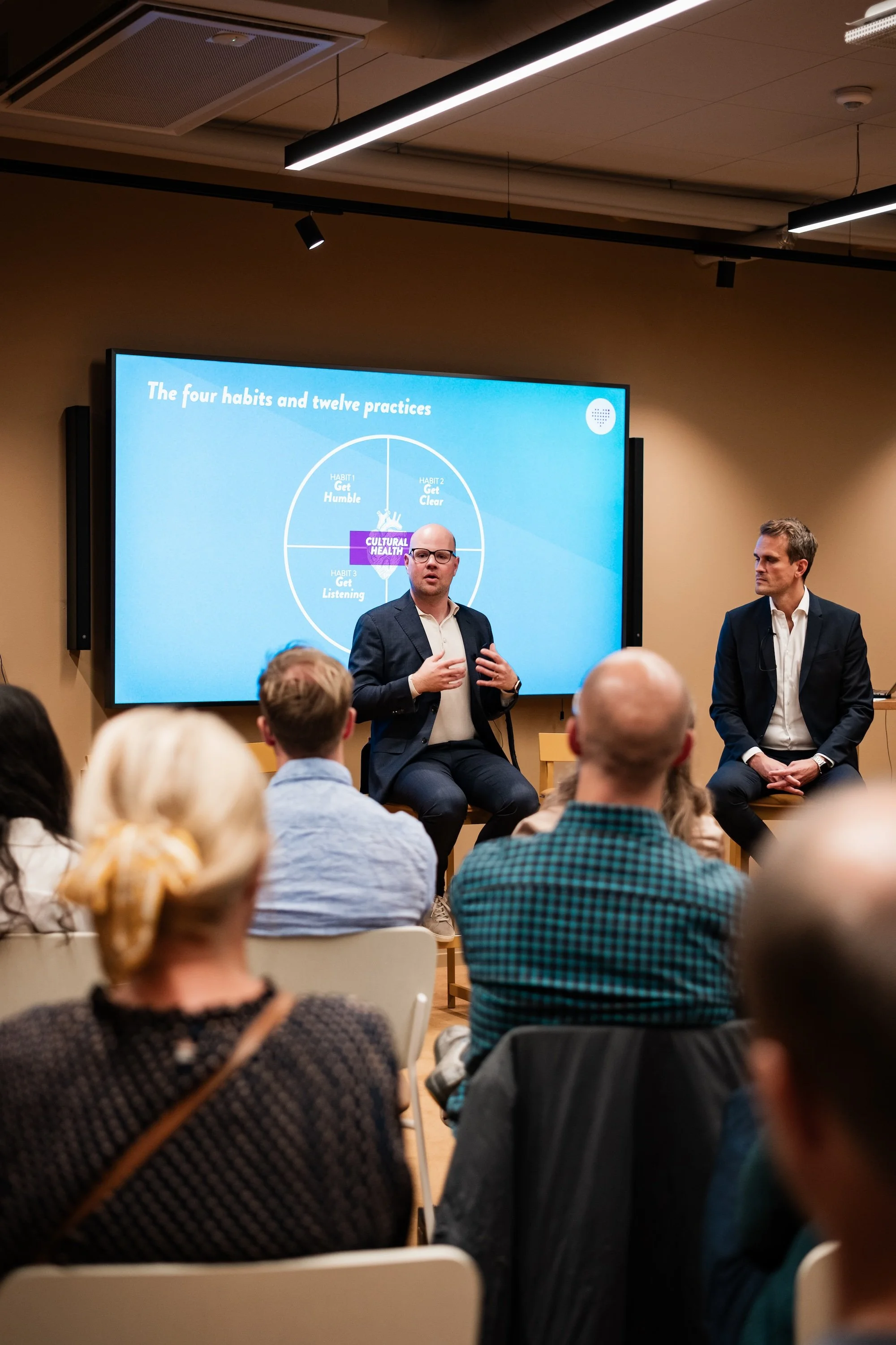 Tobias Sturesson speaking to an audience in a conference room, with a large screen behind him displaying a presentation titled 'The four habits and twelve practices' about cultural health.