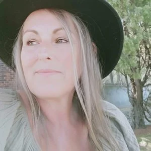 A woman with long blonde hair wearing a black hat and light-colored top, outdoors with trees and a brick building in the background.