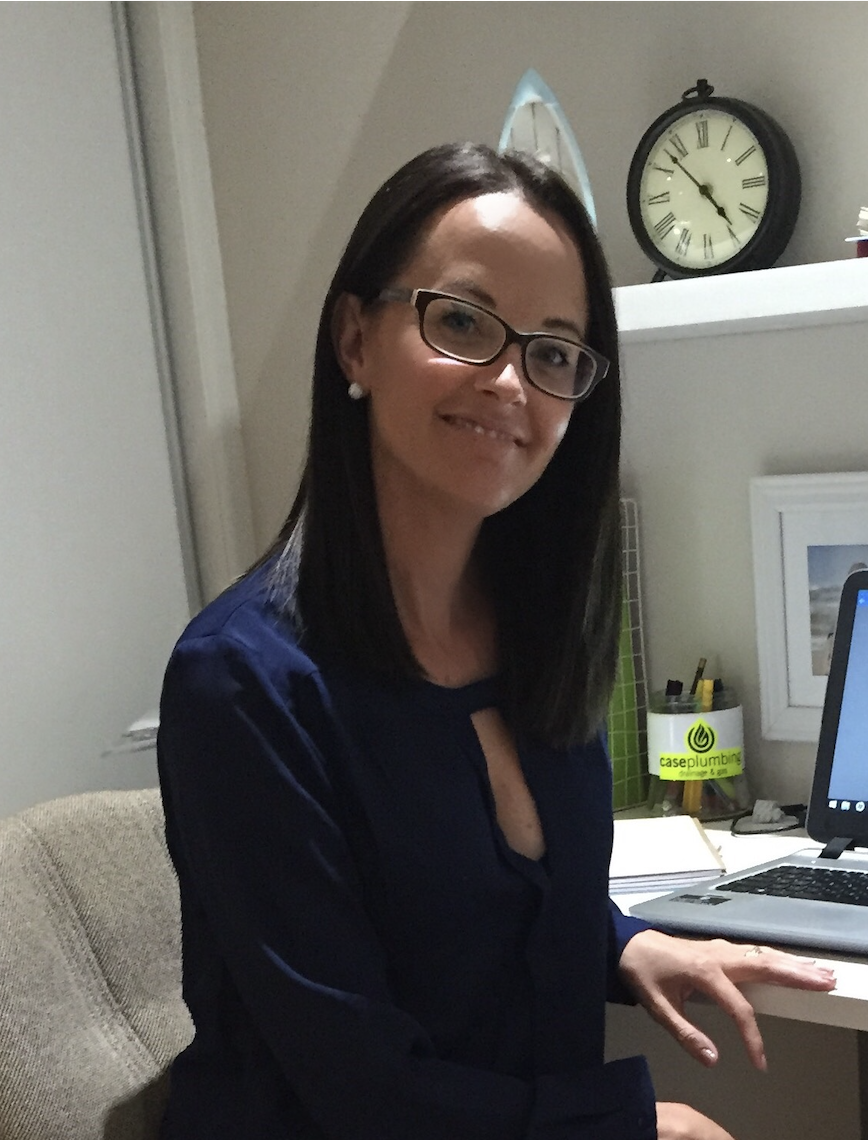 A woman with dark hair, glasses, and pearl earrings sitting at a desk with a laptop, a clock, and office supplies in the background.