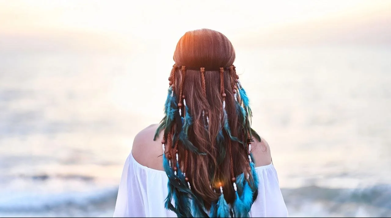 Junge Frau mit langem, buntem Haar und Perlen, trägt ein off-Shoulder-Bluse, blickt aufs Meer bei Sonnenuntergang.