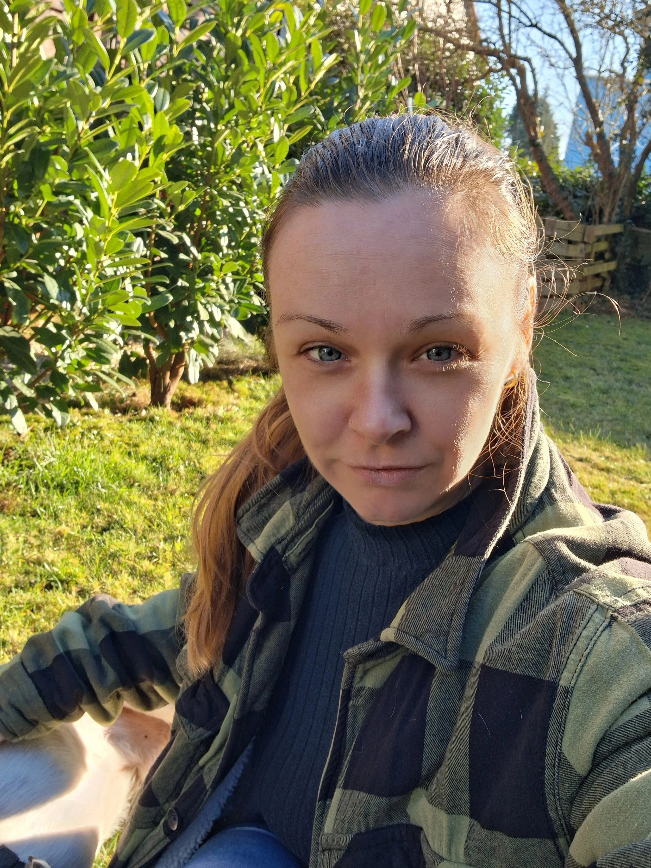 Junge Frau macht Selfie im Garten mit grüner Vegetation im Hintergrund, Sonnenlicht auf dem Gesicht, trägt Camouflage-Jacke und dunklen Pullover.
