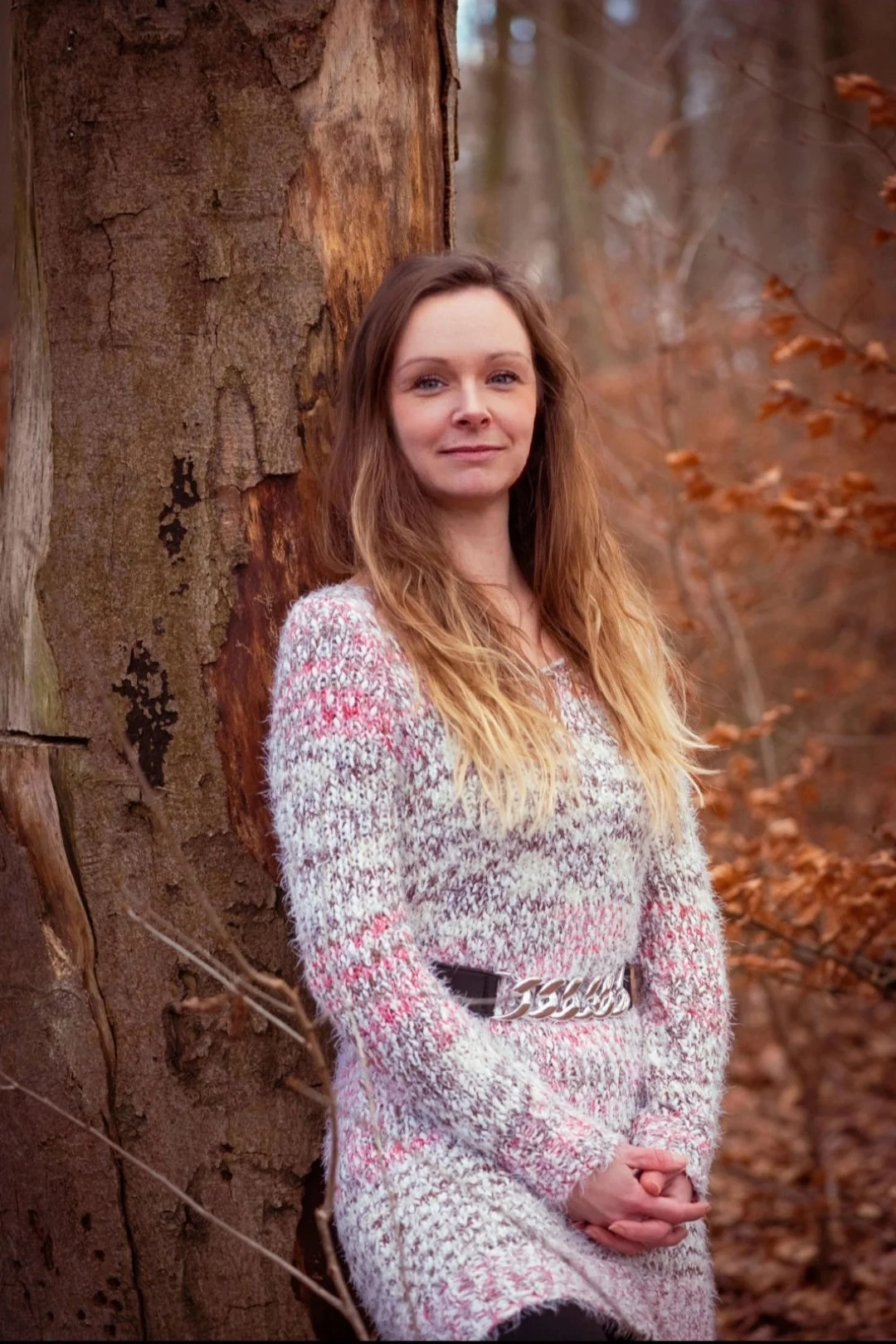 Eine Frau steht in einem Laubwald, lehnt an einem Baumstamm und schaut in die Kamera. Sie trägt einen hellen Pullover mit Muster und einen schwarzen Gürtel.