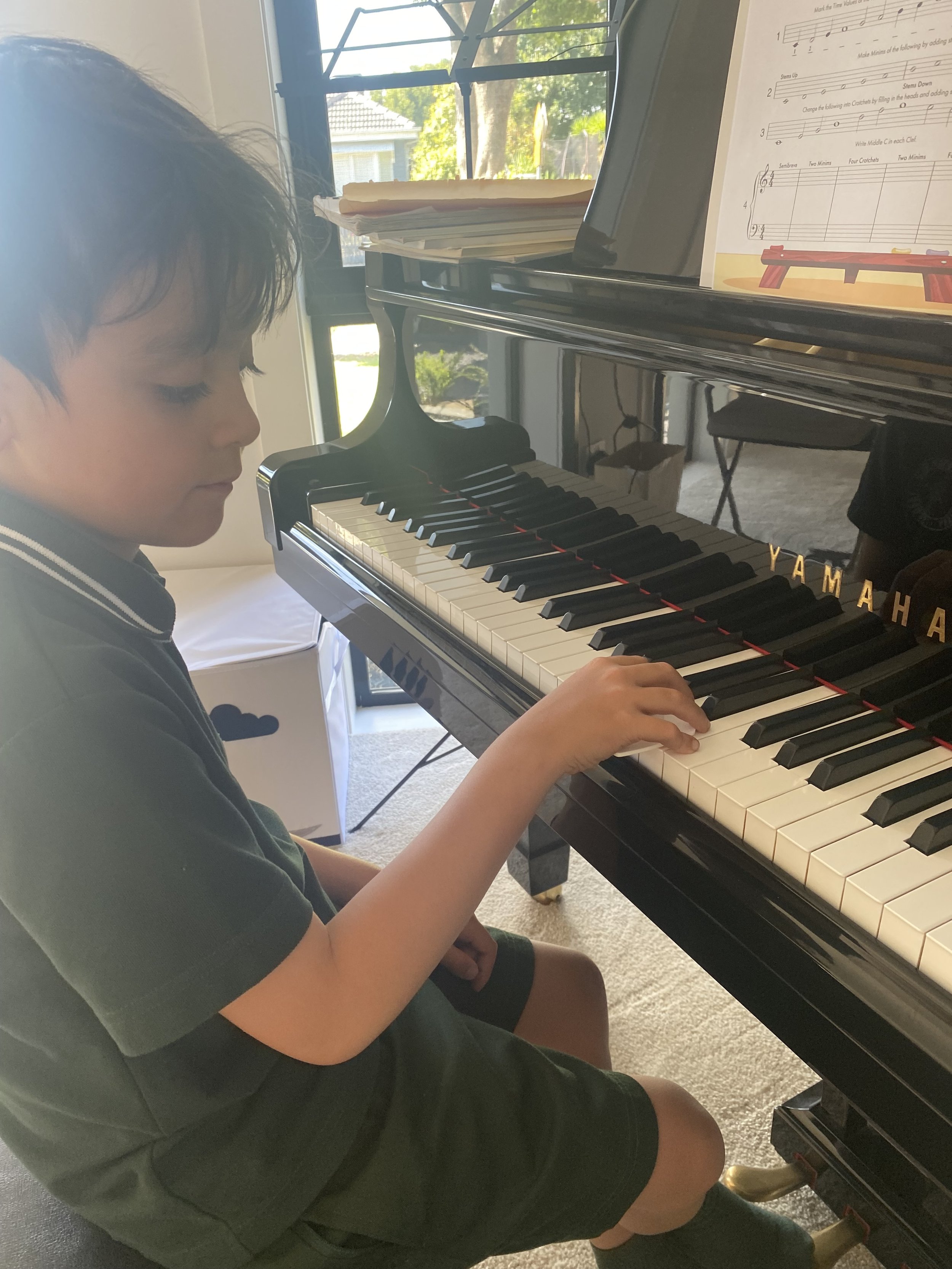 A young boy with short dark hair and a school uniform sitting at a black Yamaha grand piano, playing its white keys, with sheet music on the music stand, in a room with a window revealing trees outside.