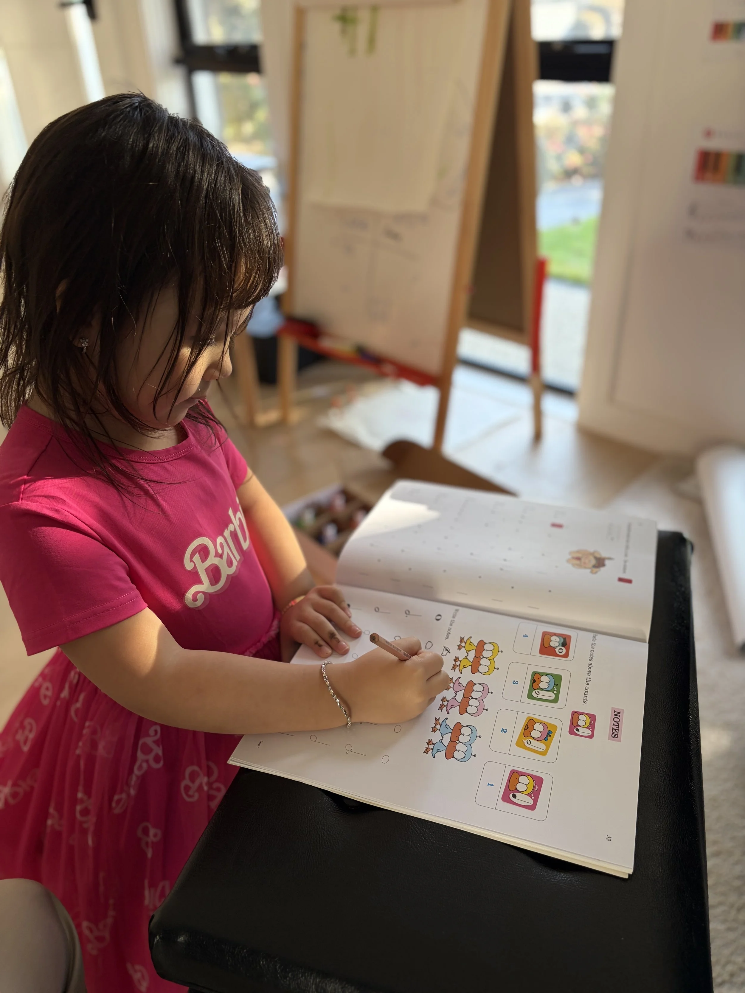 A young girl in a pink dress is writing or coloring in a workbook with pictures of colorful cartoon animals at a desk.