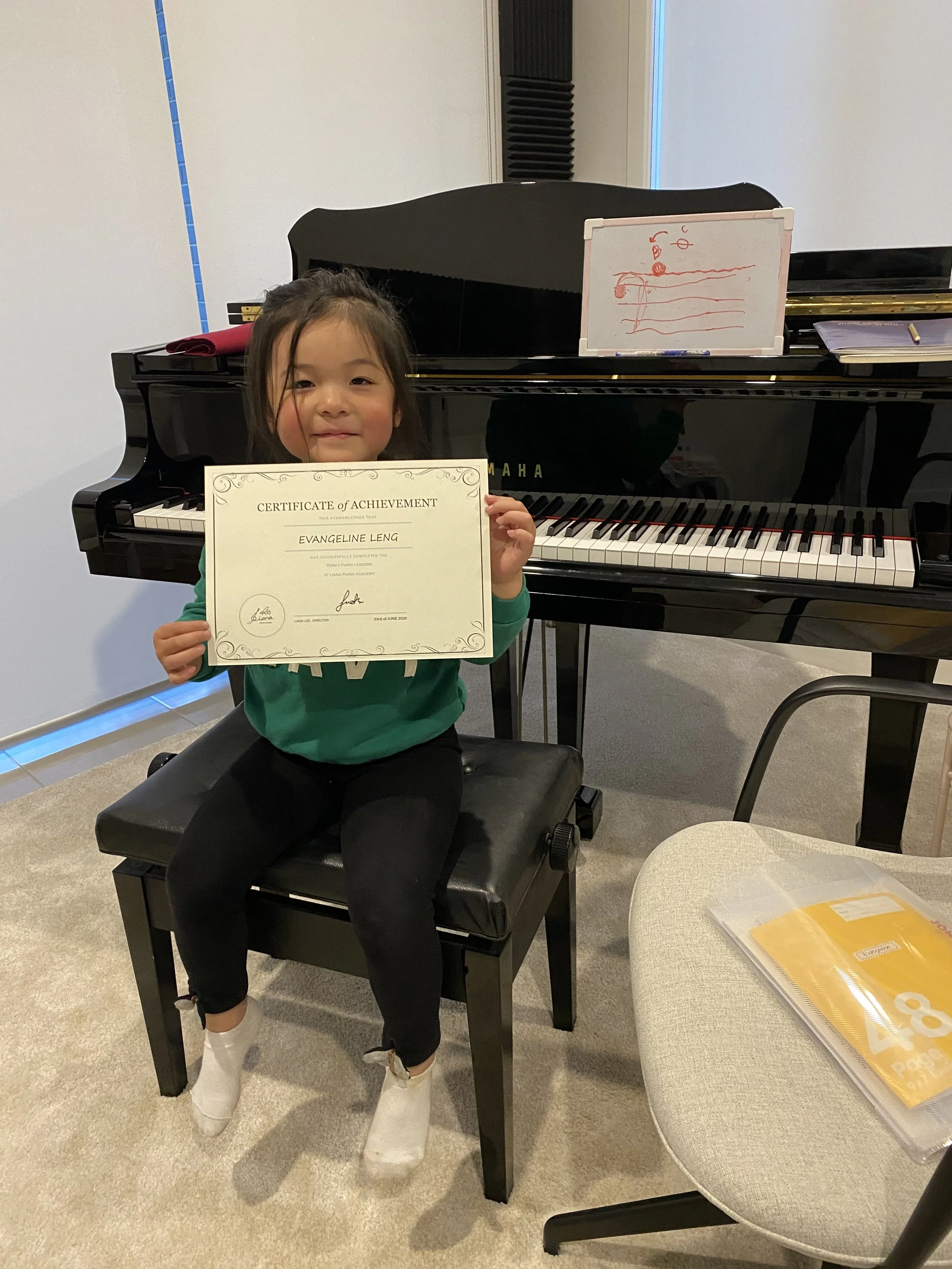 A young girl sitting on a bench in a music room, holding a certificate of achievement in front of a black grand piano with a music staff and notes drawn on a small whiteboard placed on top.