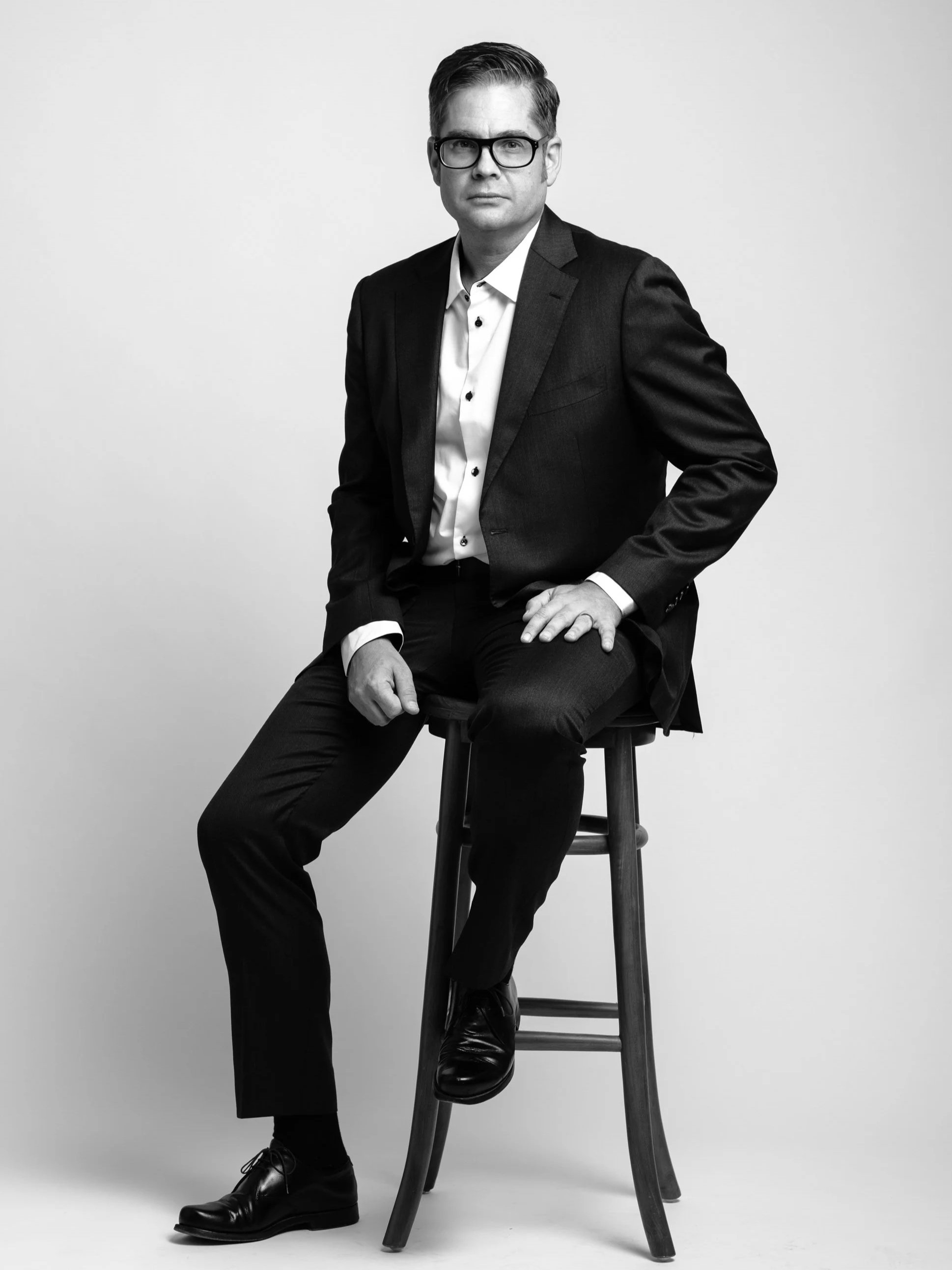 Black and white portrait of a man in a dark suit, white shirt, and black glasses, sitting on a stool against a neutral background.