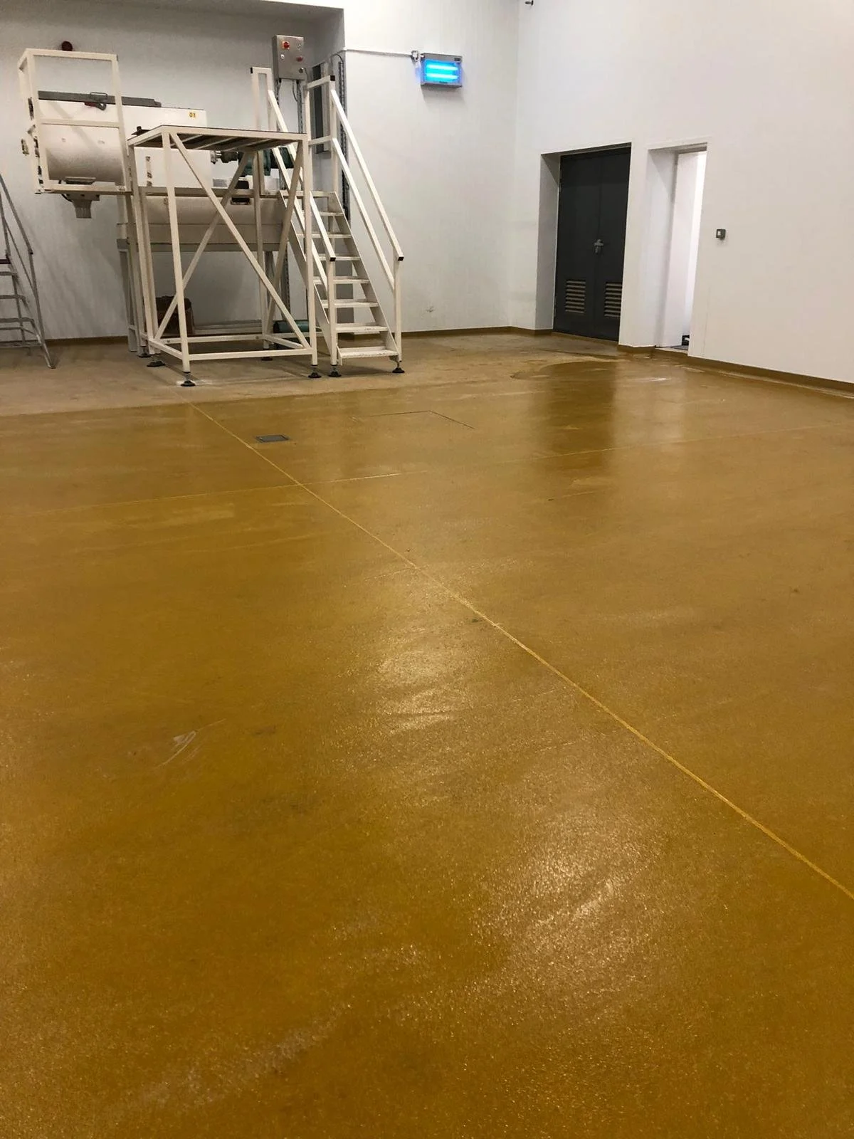 Empty indoor space with a shiny yellowish-brown floor, a white metal staircase leading to a platform, and white walls with a black door and a blue emergency exit sign.