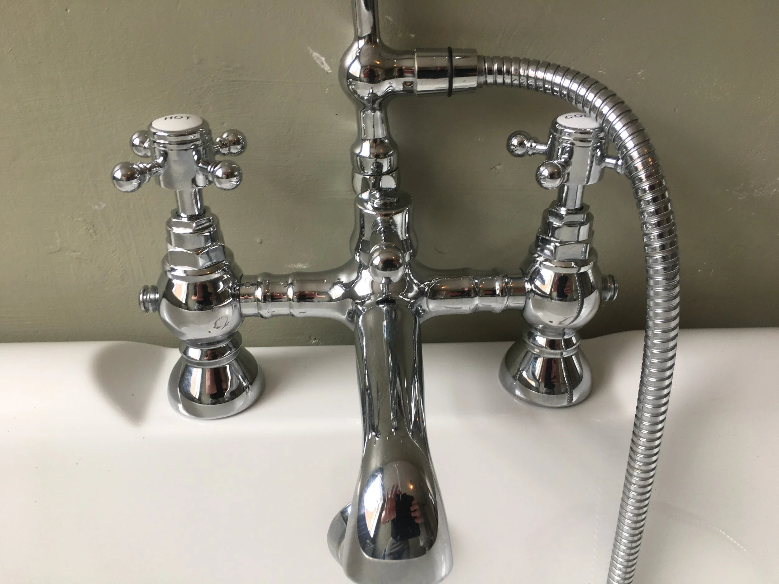 Close-up of a vintage chrome bathtub faucet with two cross handles labeled HOT and COLD, attached to a wall with a flexible shower hose.