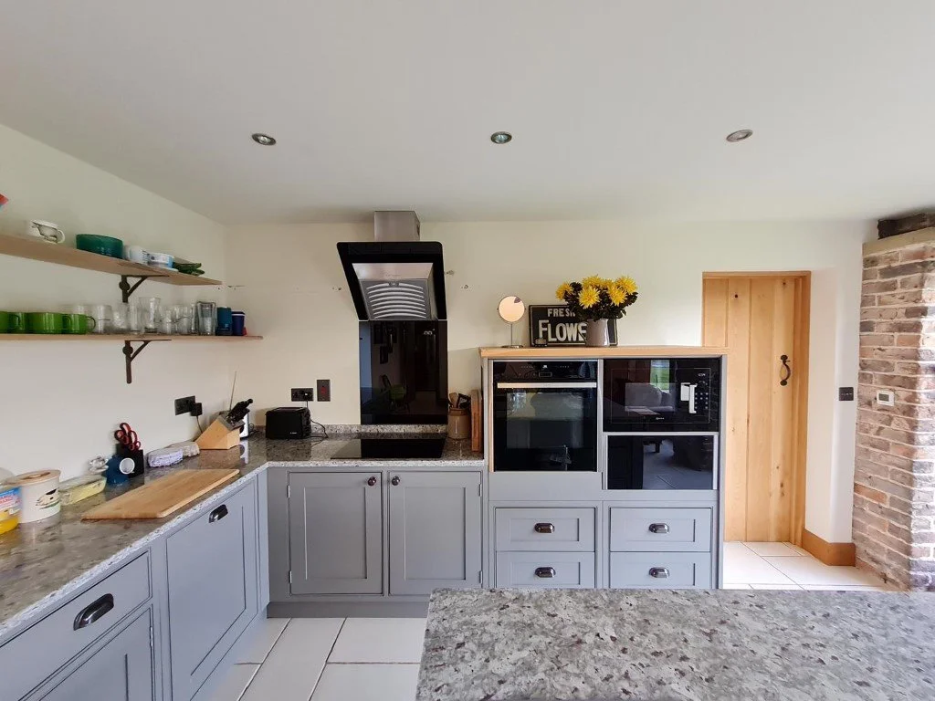 Kitchen with grey cabinets, granite countertops, open shelves holding dishes, a black range hood, and a cabinet with built-in oven and microwave. There is a wooden door on the right and a brick wall on the far right. Decorative items include a sign that reads "Fresh Flowers" and a vase with yellow flowers.
