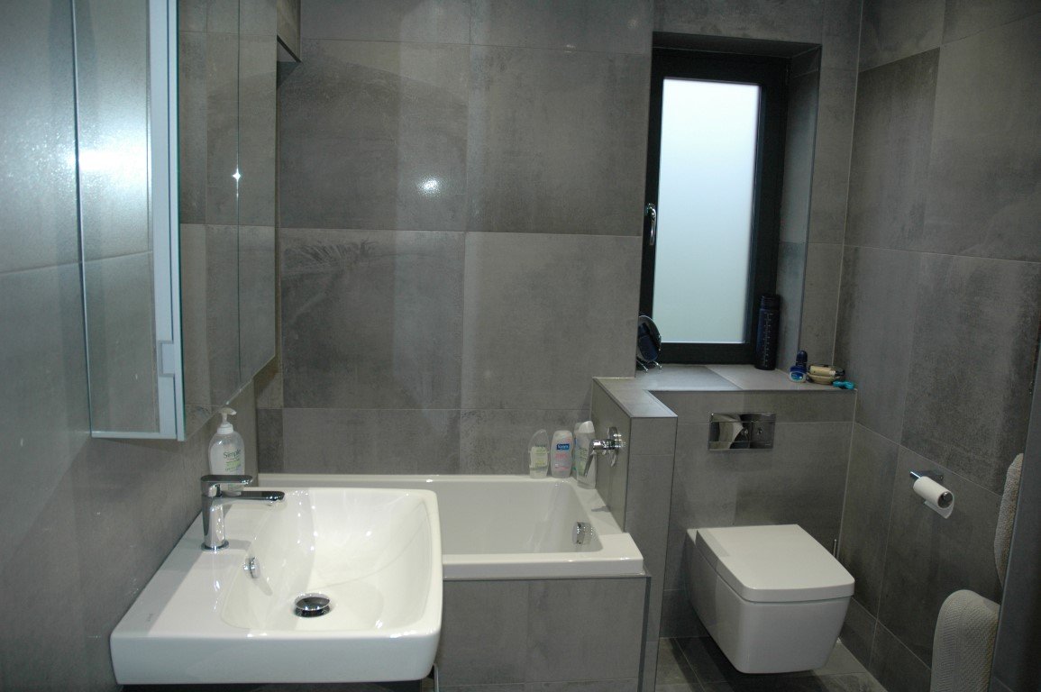 Modern bathroom with gray tiled walls, a white sink, a small bathtub, a wall-mounted toilet, a frosted window, and bathroom essentials on the window sill.