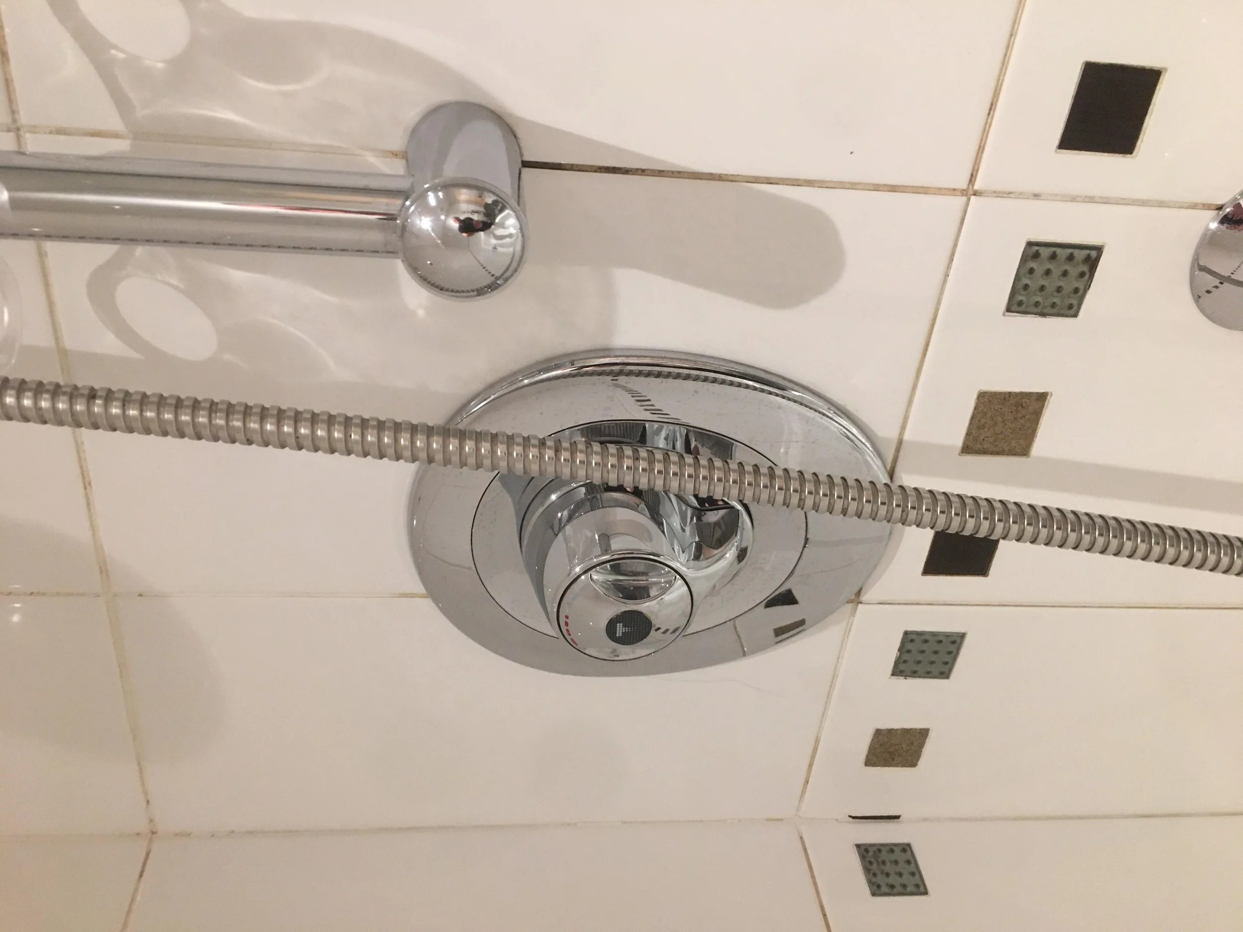 Close-up of a modern chrome shower control with a round dial, attached to a chrome shower hose and a handheld showerhead on a tiled wall with decorative square tiles.