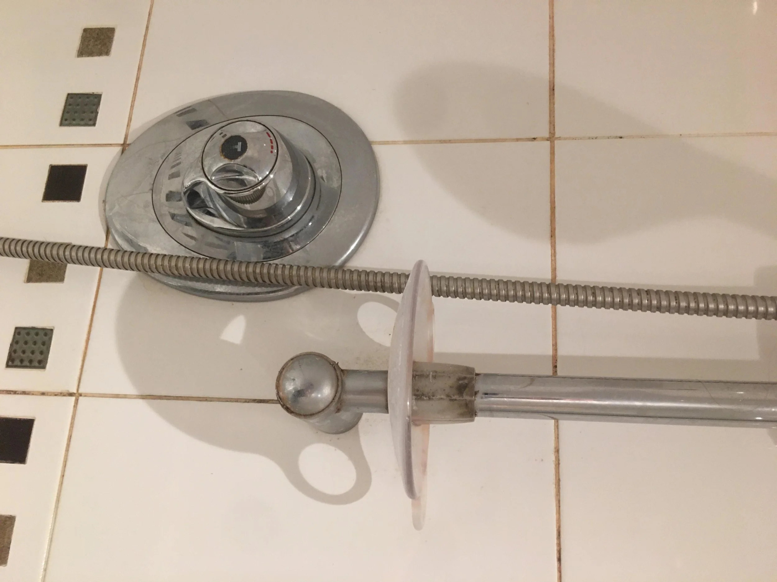 A close-up of a shower control panel with a round knob set to the warm water setting, mounted on a tiled wall with a shower head and hose below it.