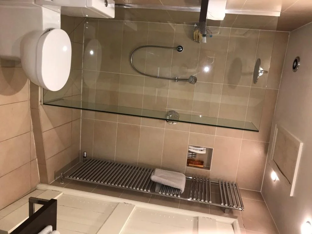 Bathroom with a toilet, a glass-enclosed shower area with a handheld showerhead, towel rack, and beige tiled walls and floor.