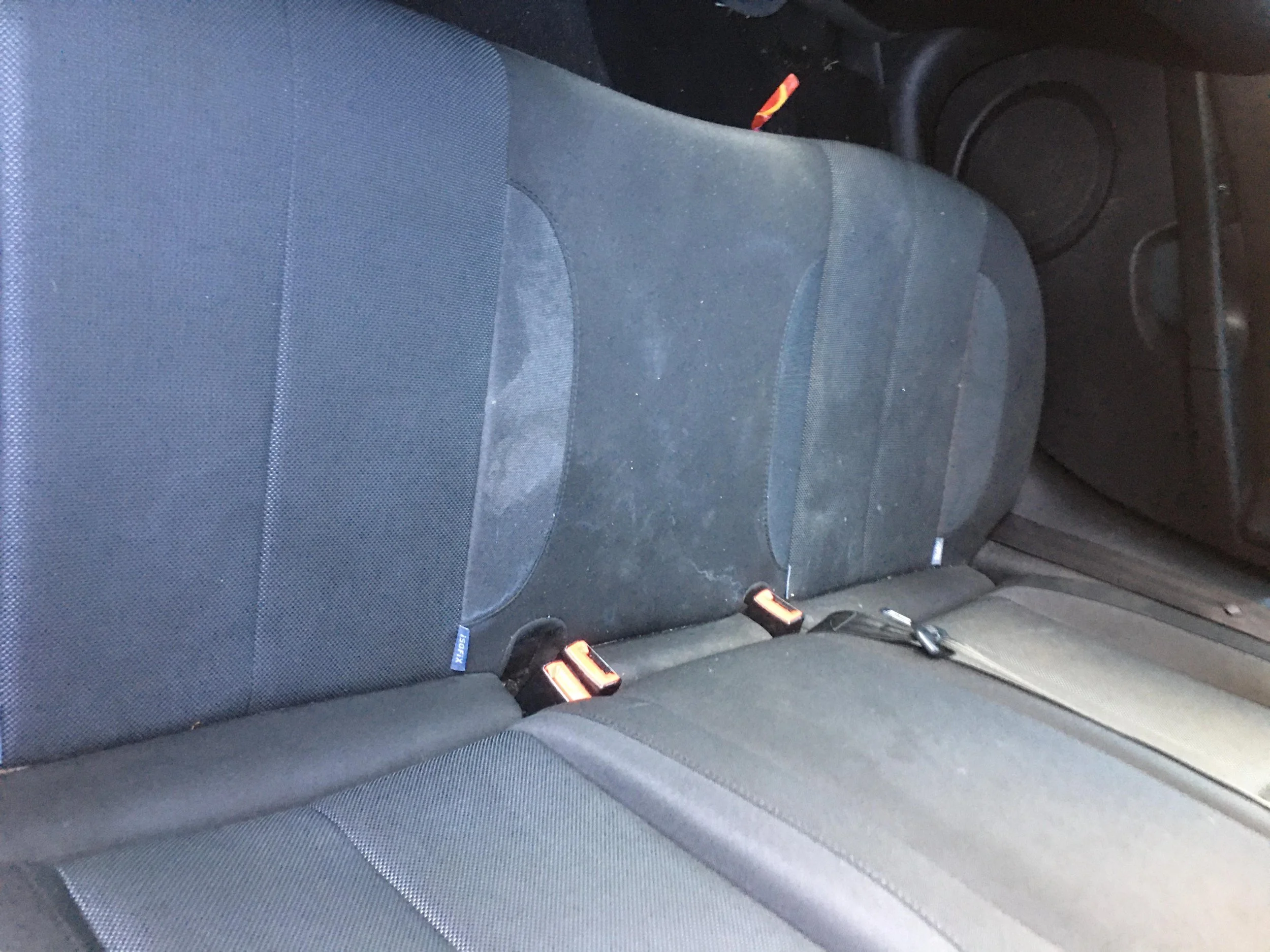 Back seat of a car with black fabric upholstery, some dust and dirt visible, and seat belts.