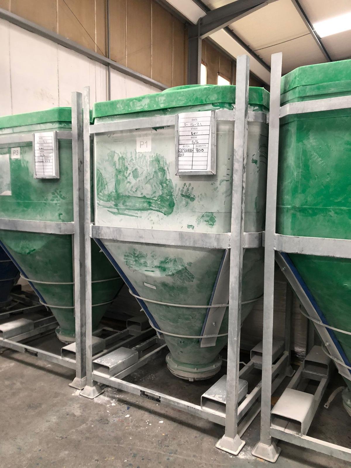 Three large green storage hoppers with metal frames, used for industrial purposes, labeled with handwritten signs, inside a warehouse.