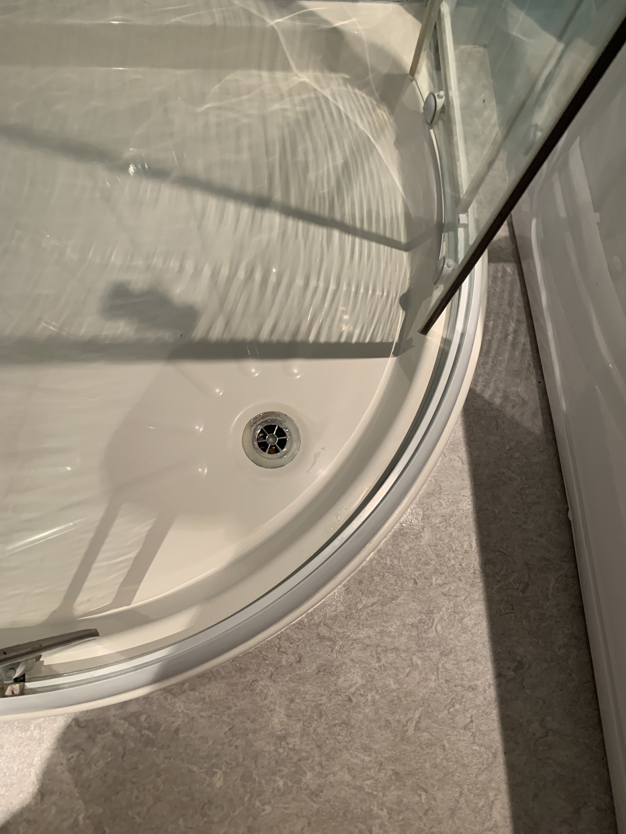 Close-up view of a shower with a curved glass door and a white acrylic shower base with a drain, placed on a beige tiled floor.