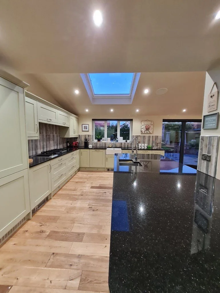 Modern kitchen with cream-colored cabinets, black countertop, wooden flooring, a skylight, and large windows leading to an outdoor area.