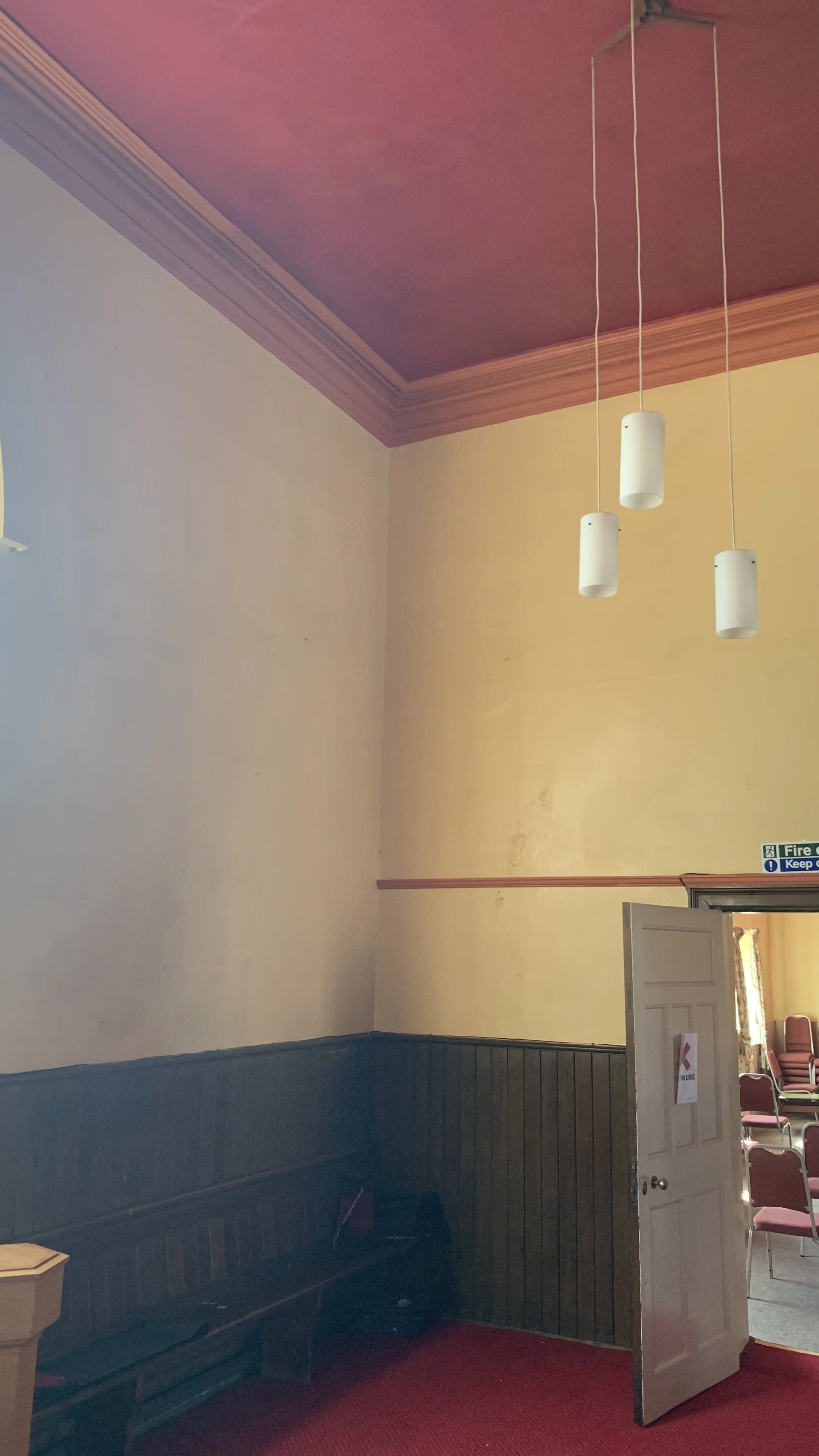 Interior view of a room with cream-colored walls and a pink ceiling, featuring three hanging white pendant lights. Partially open door reveals chairs inside, and a fire safety sign is visible on the wall.