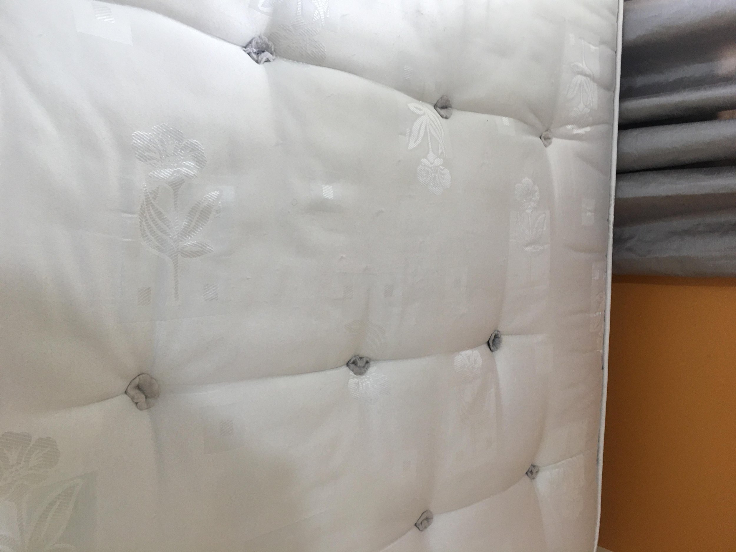 Close-up of a cream-colored, tufted mattress with floral patterns and gray fabric corner, with gray curtains and a yellow wall in the background.