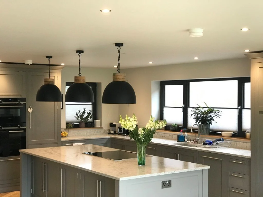 Modern kitchen with gray cabinets, marble countertops, three black pendant lights, large window with plants, and a vase of white flowers on the island.
