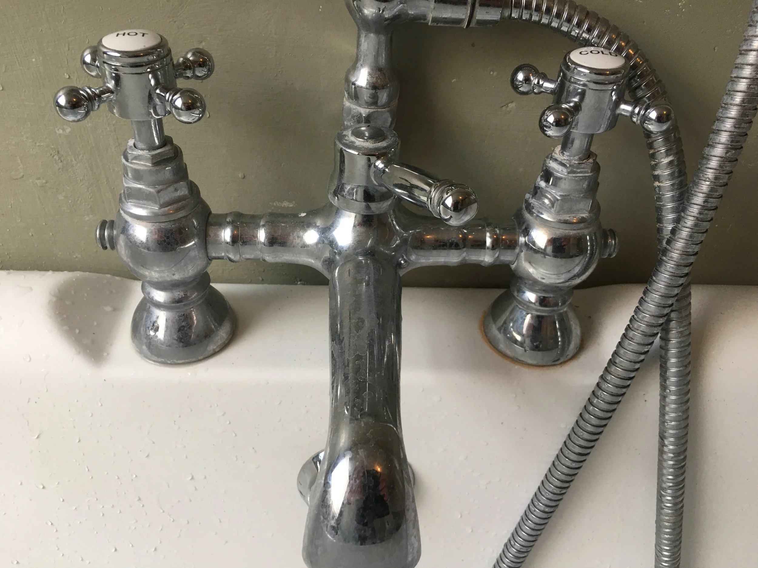 Close-up of vintage chrome faucet with separate hot and cold handles, mounted above a white sink with water drops, against a greenish wall.