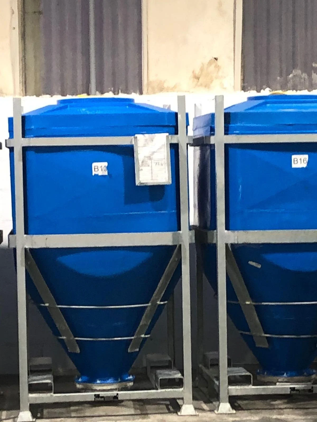 Two large blue industrial hoppers with metallic frames positioned in front of a wooden wall.