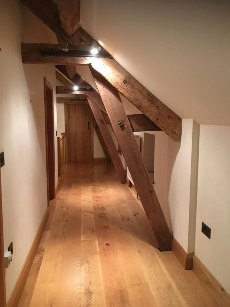 A hallway with wooden flooring and wooden beams on the ceiling. Large wooden beams are supported by angled wooden braces, creating an architectural feature in the space. The hallway is lit by small ceiling spotlights.