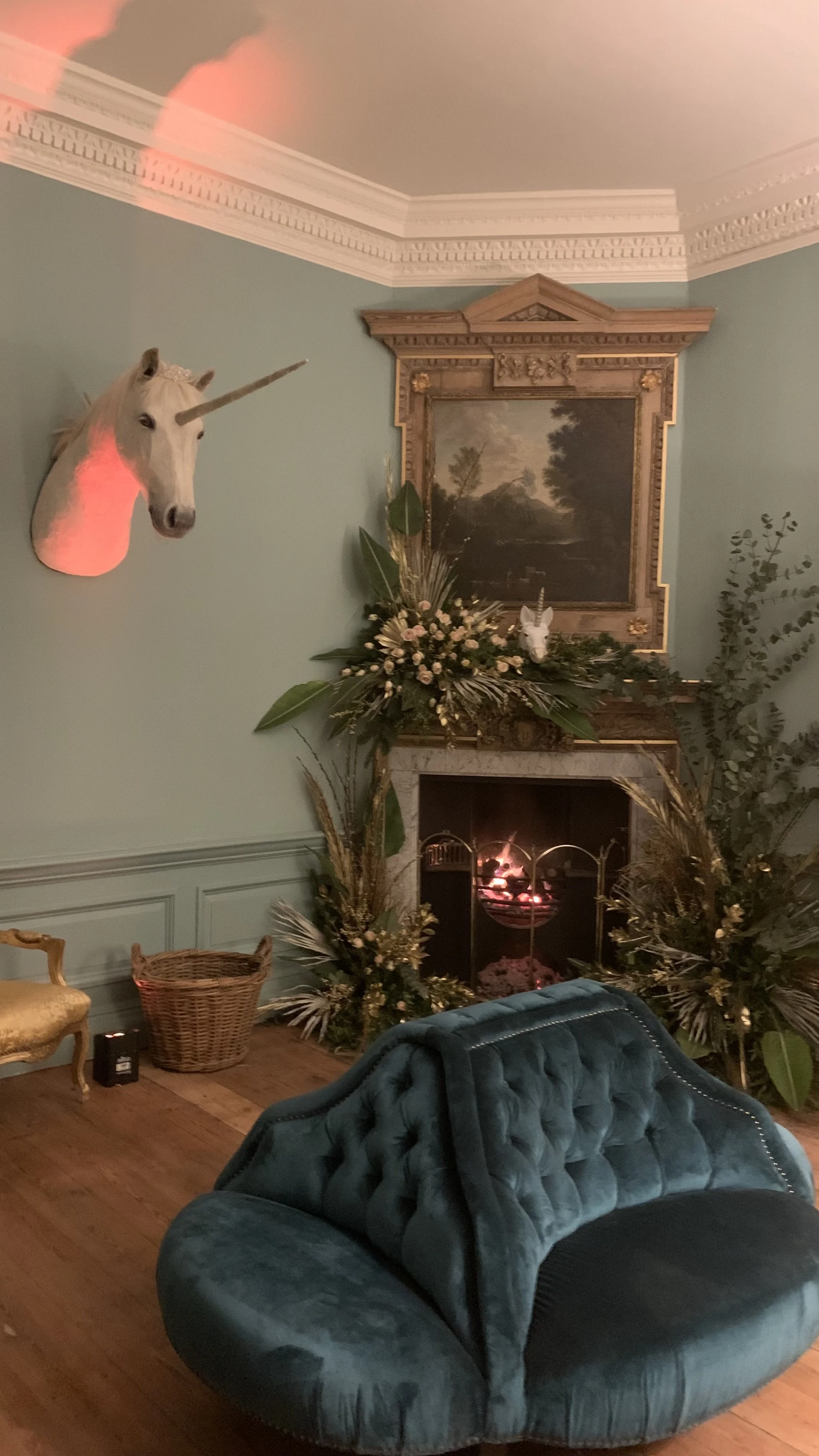 A living room with a fireplace decorated with flowers and greenery, a portrait above it, a sword and shield, and a unicorn head mounted on the wall. There is a blue velvet sofa, a gold chair, and a wicker basket.