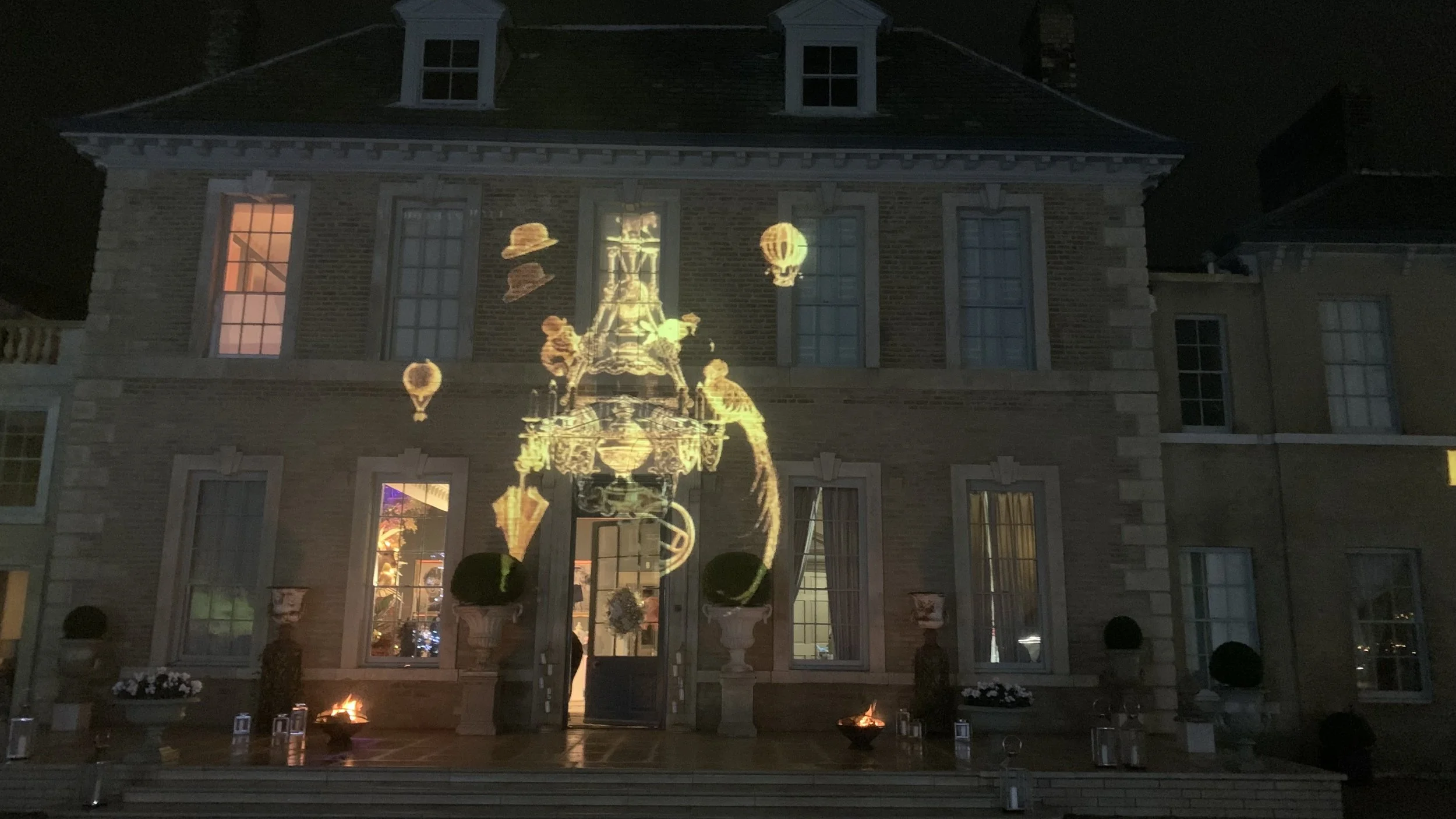 A projection of vintage travel-themed images like hot air balloons, a ship, and a hat is displayed on the exterior of a brick house at night.