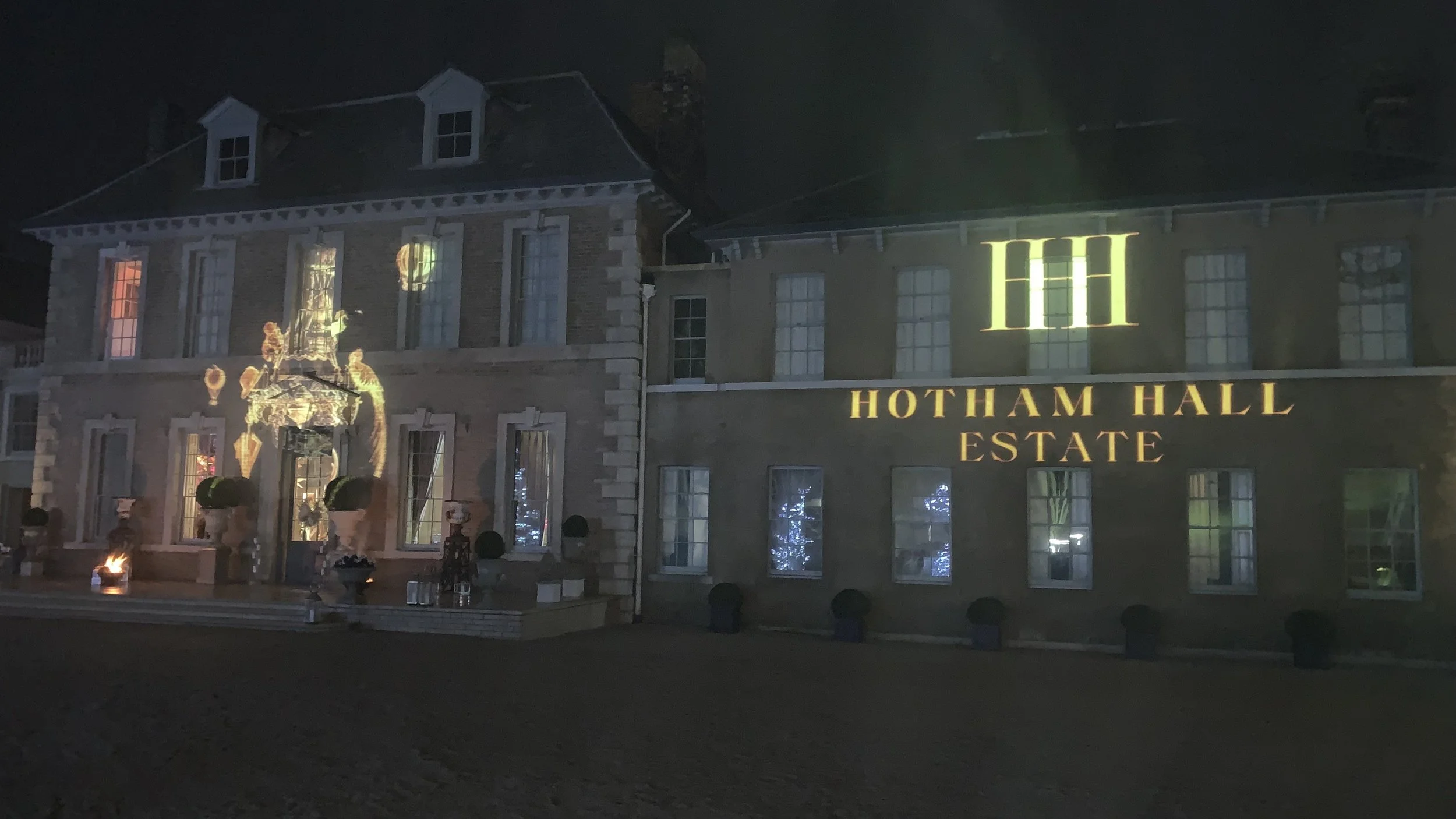 Nighttime view of Hotham Hall Estate, a large brick building with multiple windows, some with lights on. The estate's name is projected in yellow light on the front of the building, and there are decorative lights and ornaments outside.