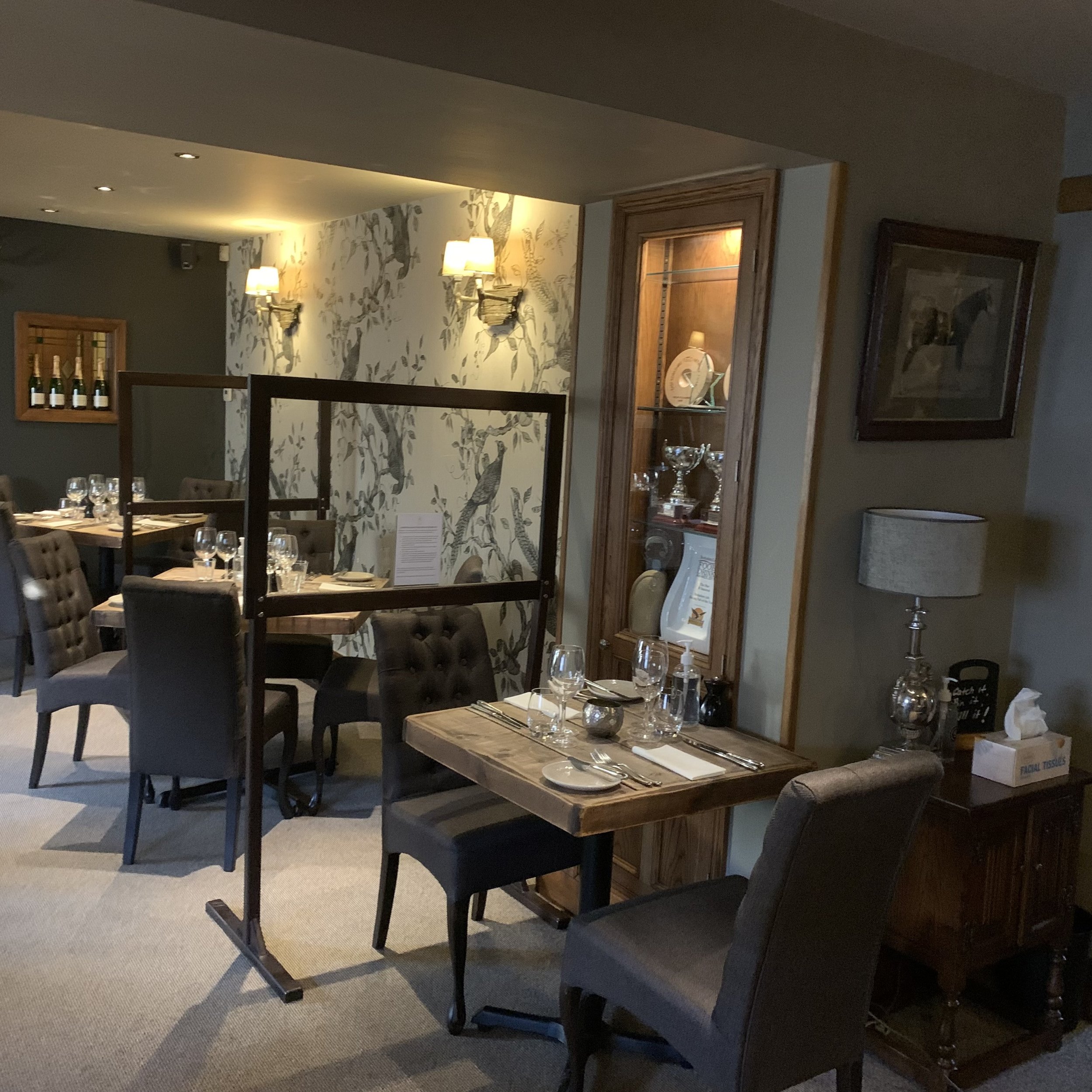 Indoor dining area of a restaurant with table settings, including wine glasses, napkins, and cutlery, surrounded by upholstered chairs. There is a wooden display cabinet with glassware and trophies, a wall with floral wallpaper, and wall-mounted lamp